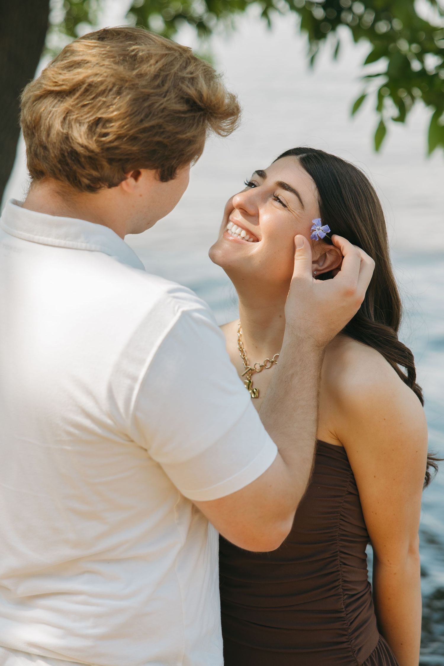 Man placing a small purple flower in smiling womans hair by a lakeside.