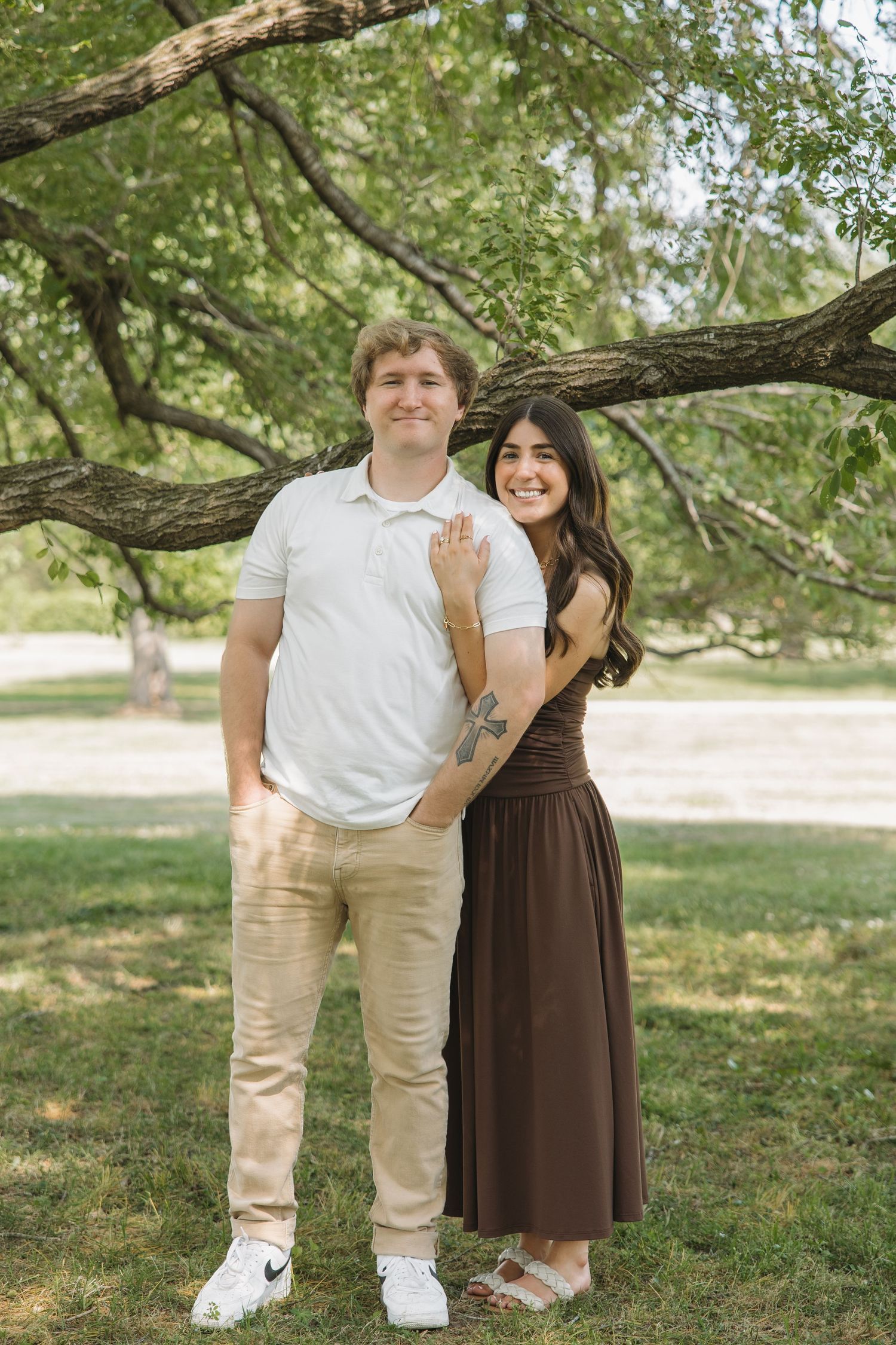 Couple posing under a tree in a park, woman in brown dress showing engagement ring, man in white polo.