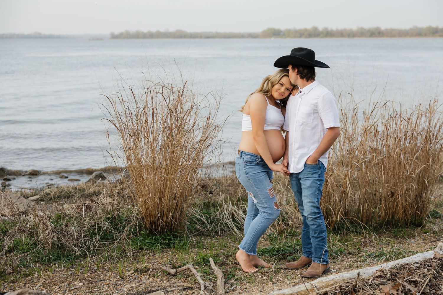 Maternity photo of couple holding hands near tall reeds by lake, man in cowboy hat admiring pregnant partner.
