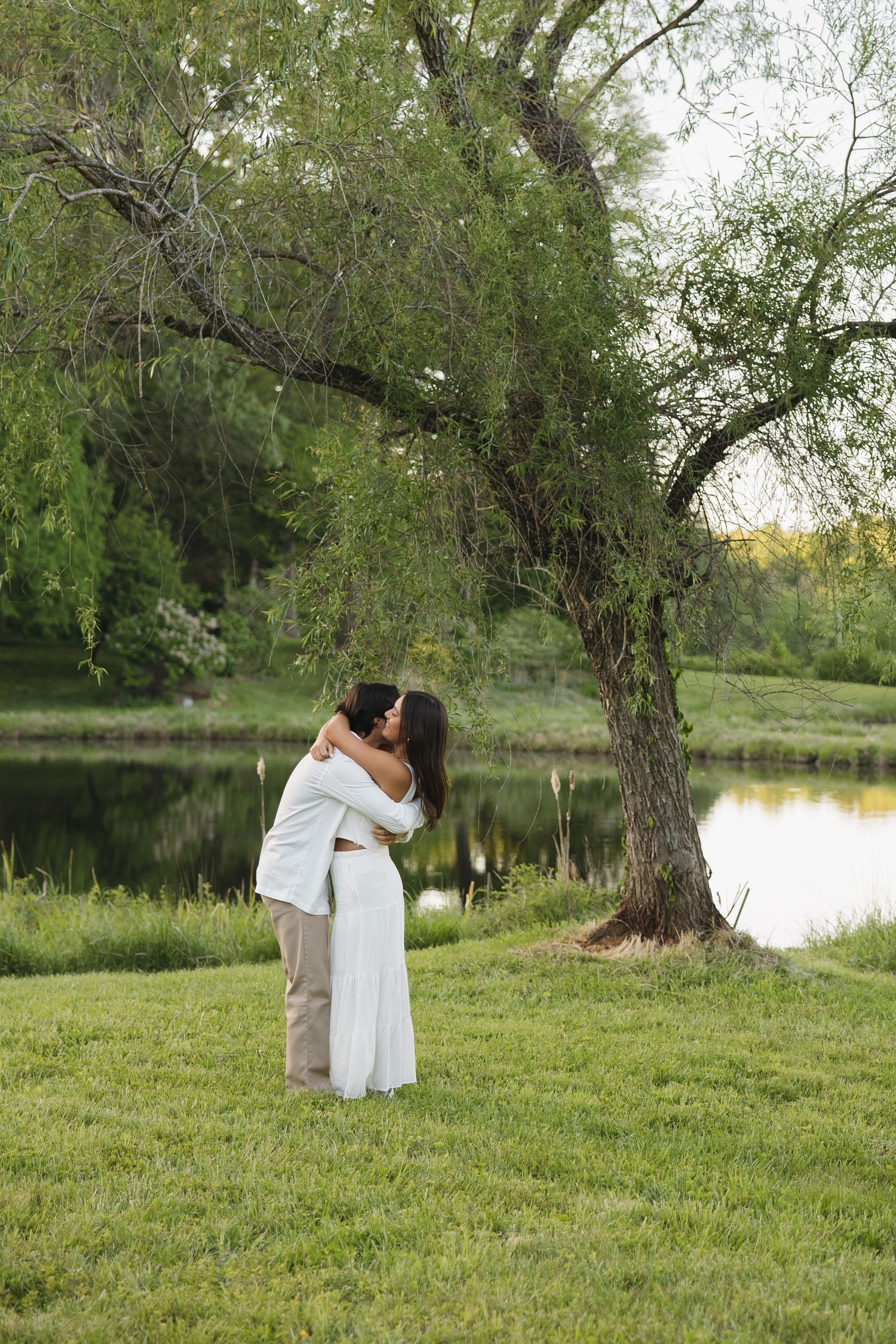 Couple embracing and kissing near a serene pond under a willow tree in a lush green park.