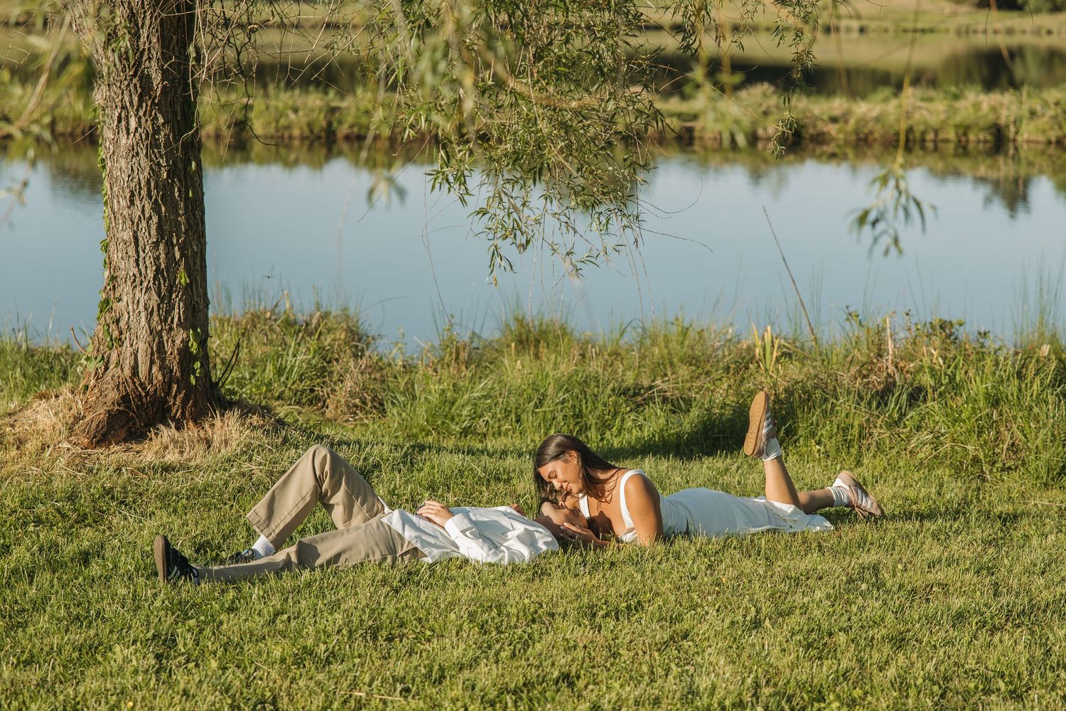 Couple lying close together on green grass beneath a tree near a peaceful reflective pond in soft sunlight.