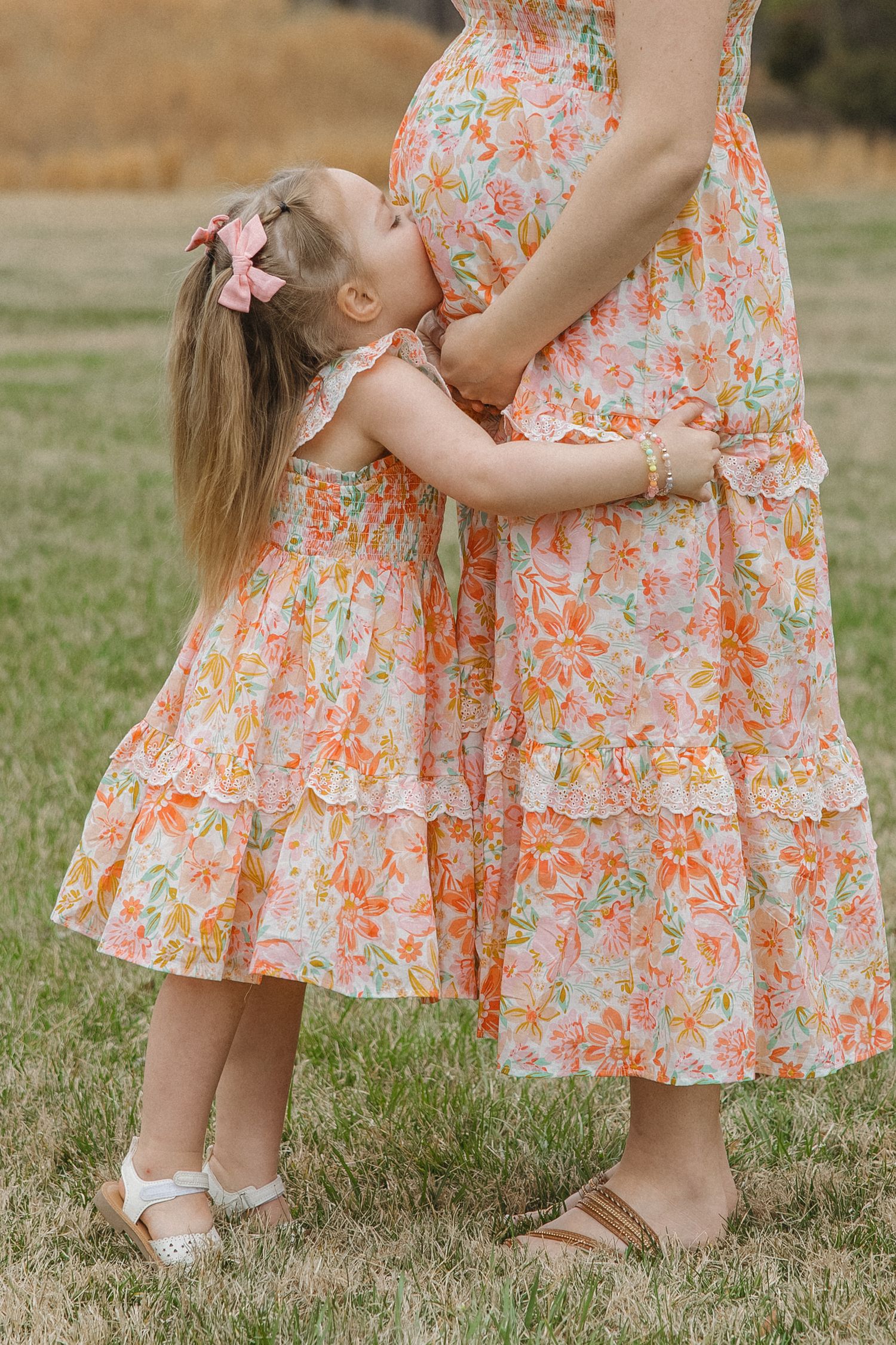 Young girl in floral dress kisses pregnant mothers belly while embracing her outdoors in a grassy field.
