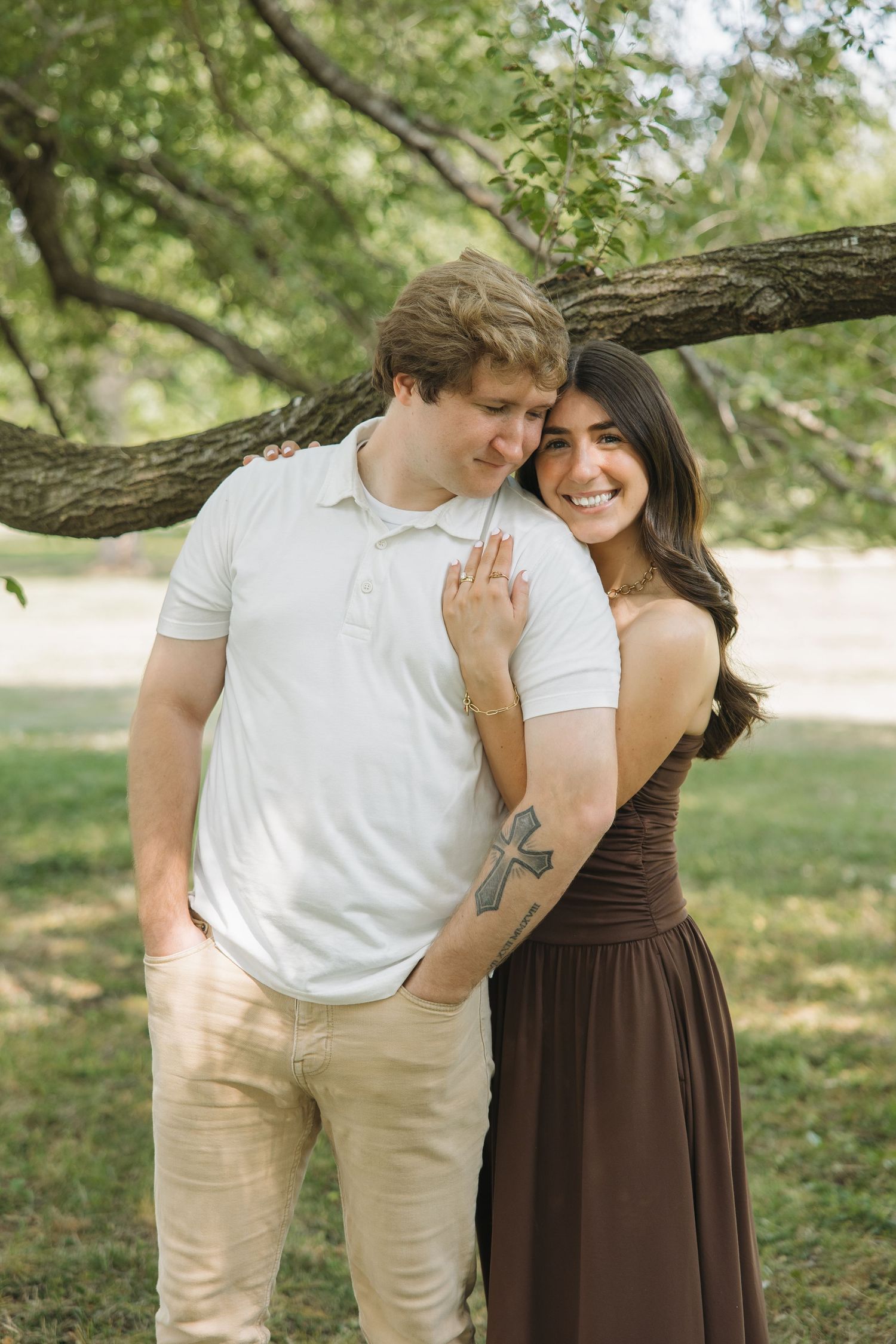 A happy couple poses under a tree, woman in brown dress with cross tattoo smiling brightly against mans shoulder.