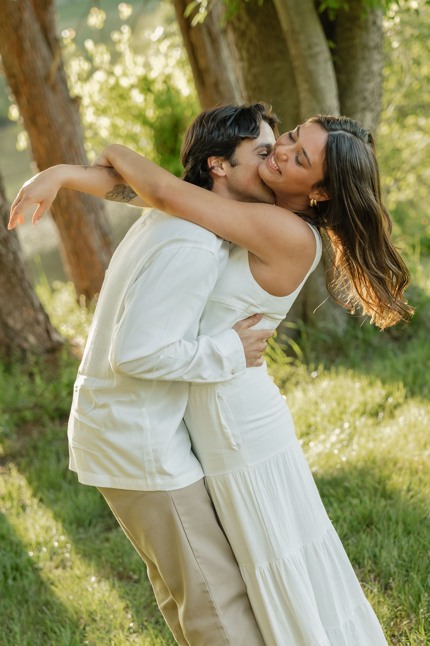 Couple kissing and embracing romantically in a sunlit forest clearing with lush green trees behind them.