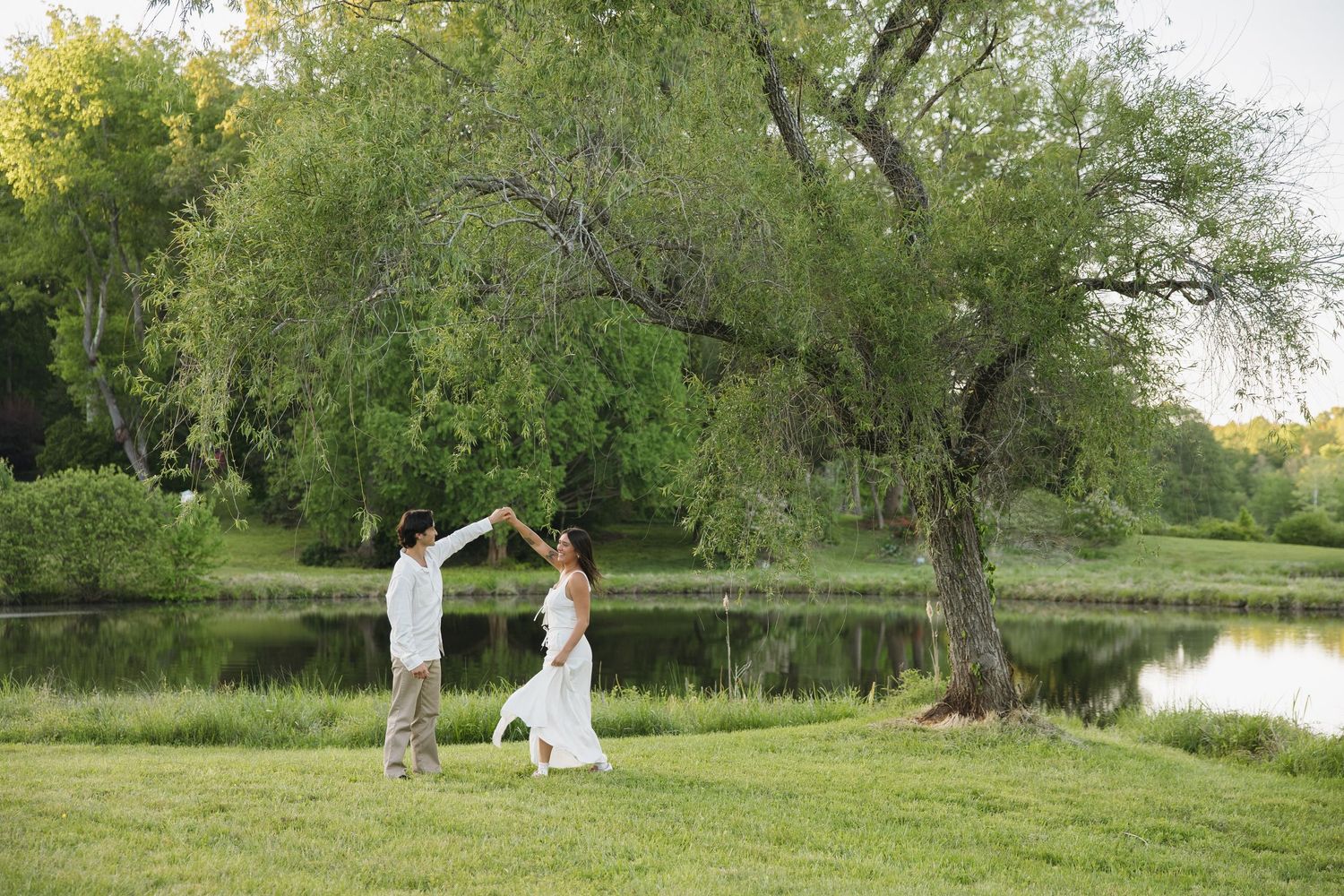 Couple dancing and twirling near a willow tree by a reflective pond in a lush green park.