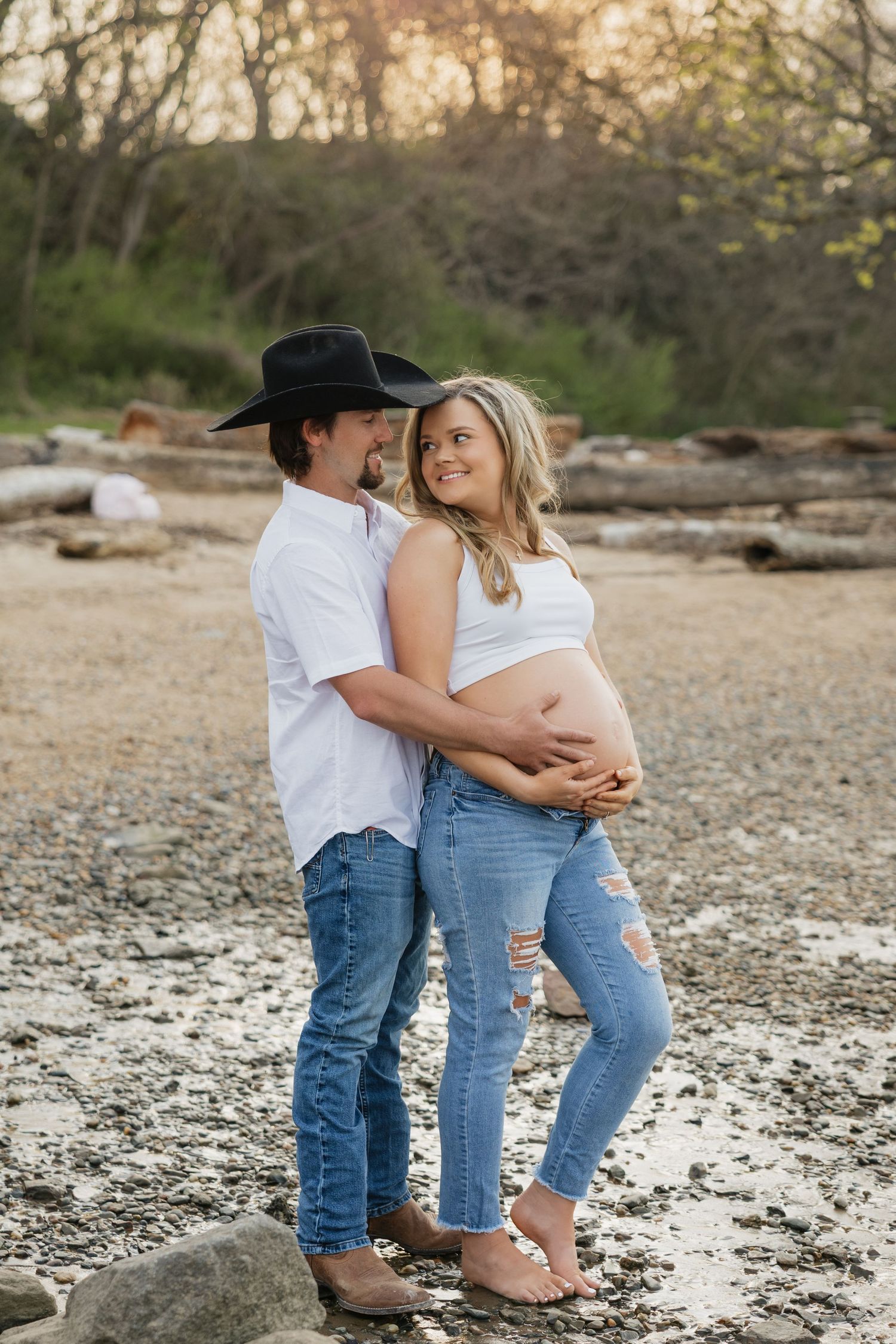 Couple embracing at golden hour on pebbly beach, pregnant woman in white crop top, man in black cowboy hat.