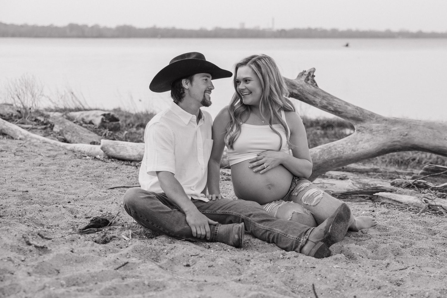 Black and white photo of couple sitting on beach by driftwood, pregnant woman and partner in cowboy hat smiling.