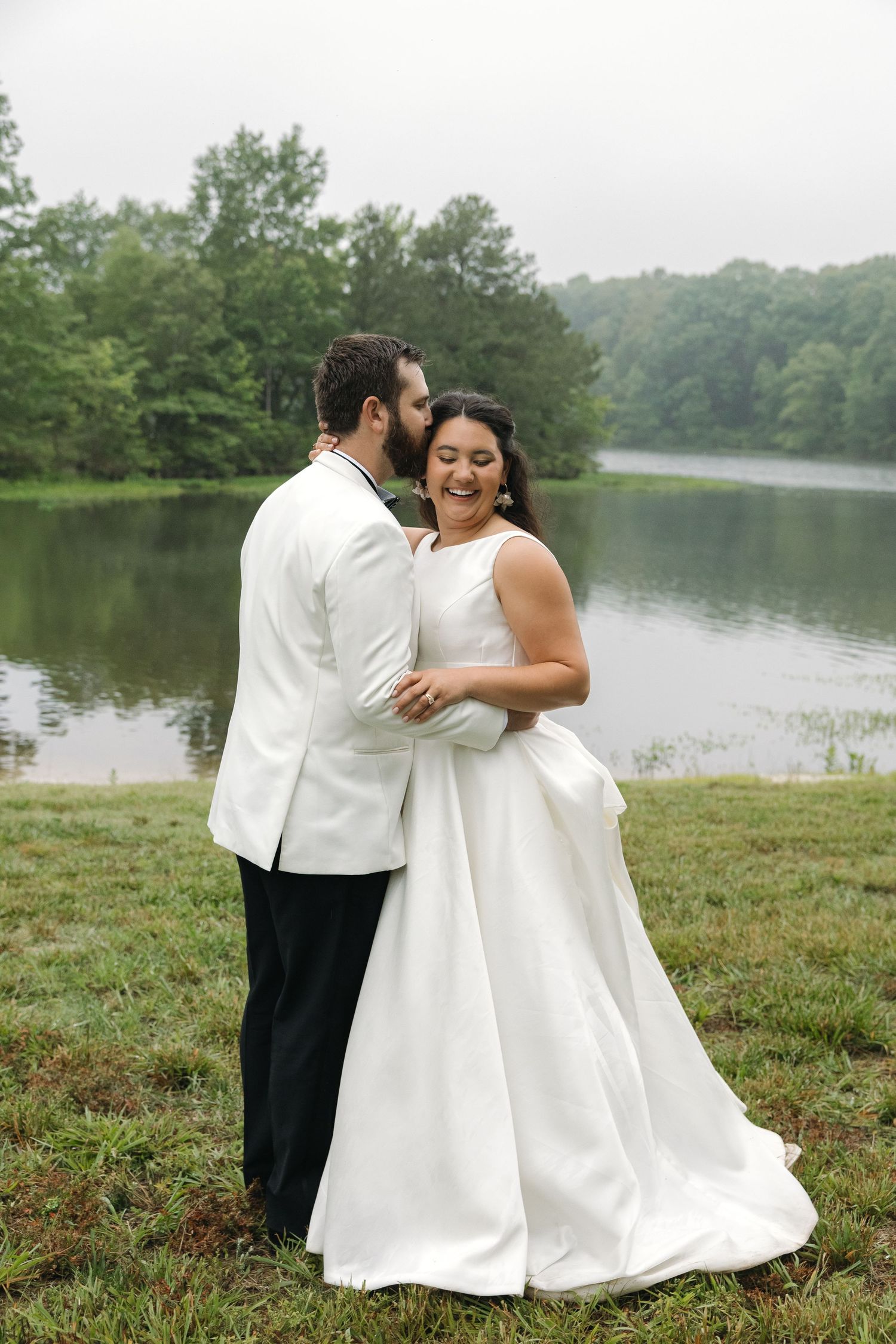 Bride laughing joyfully as groom whispers in her ear while standing together by a misty lake.