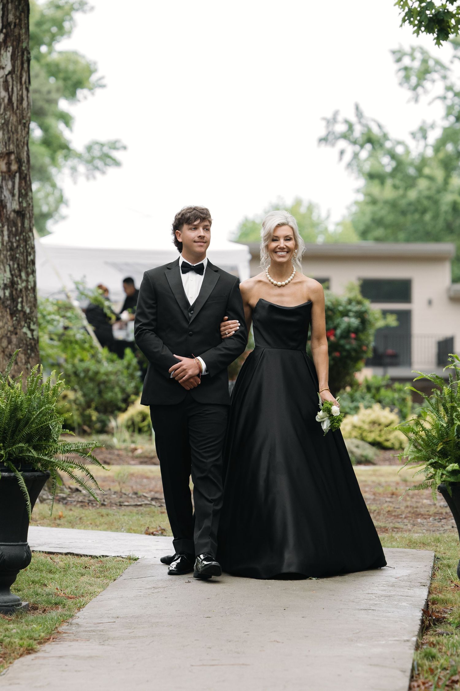 Groomsman in black tuxedo escorts bridesmaid in black strapless gown down outdoor wedding aisle.
