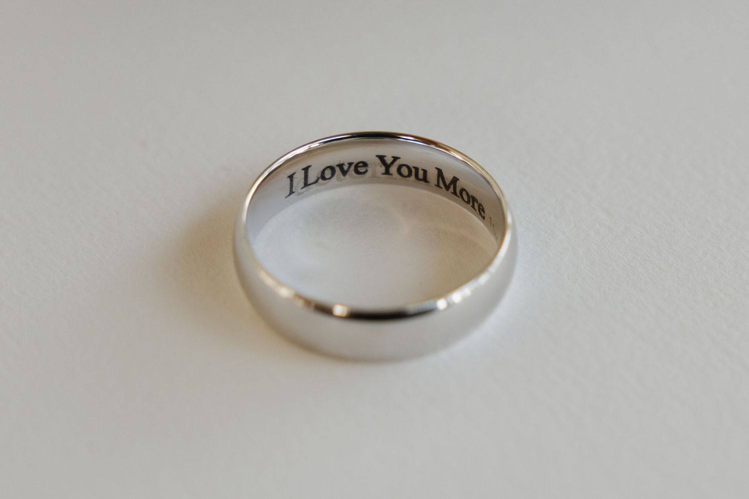 Silver wedding band with romantic engraved message I Love You More on white background close-up detail shot.