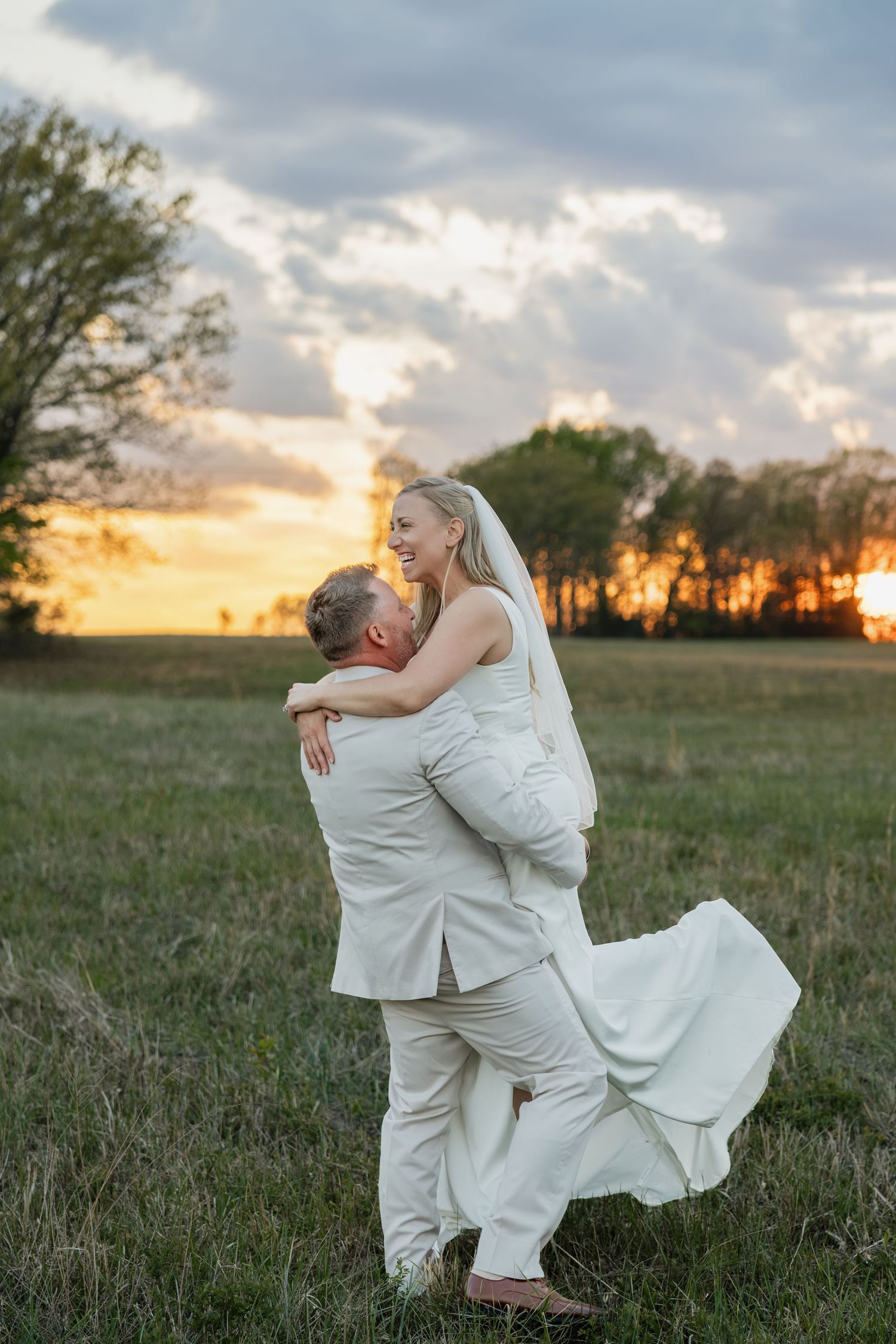 Groom lifts laughing bride in a field with a glowing orange sunset behind them.