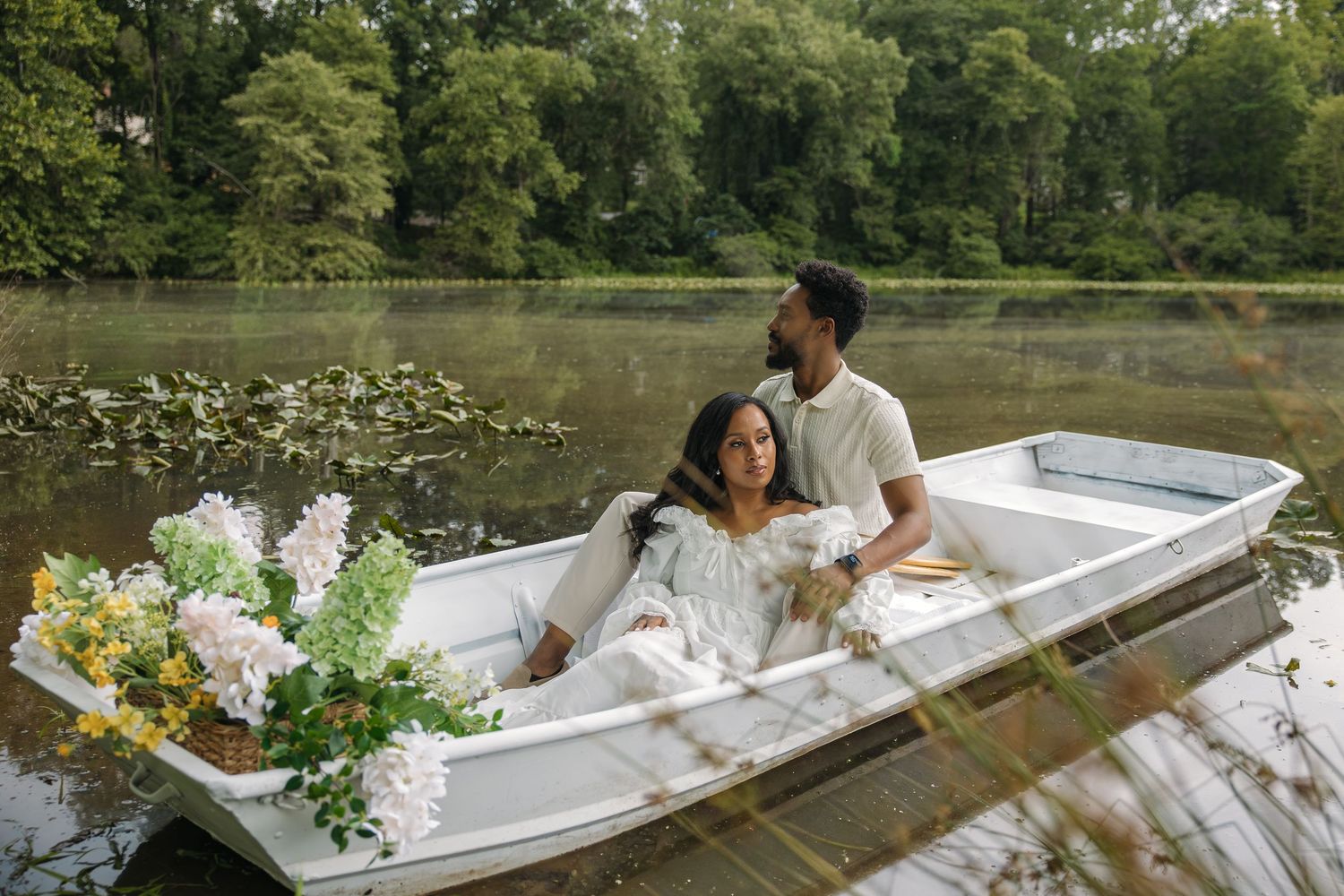 Couple relaxing in a white boat on a calm lake surrounded by lush green trees and flowers.