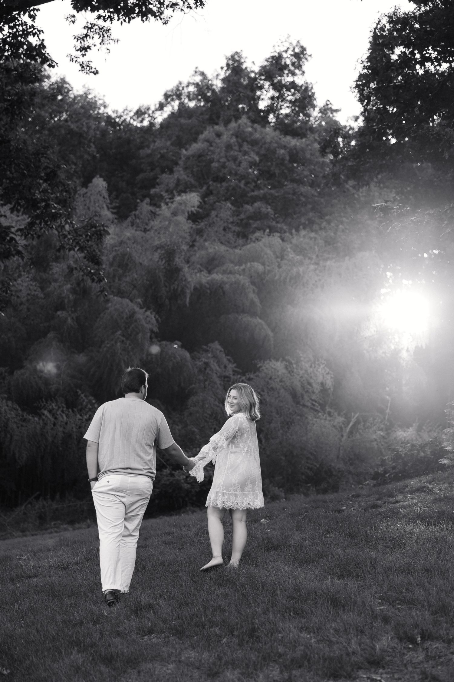 Black and white photo of a couple holding hands walking through a sunlit grassy field toward lush green trees.