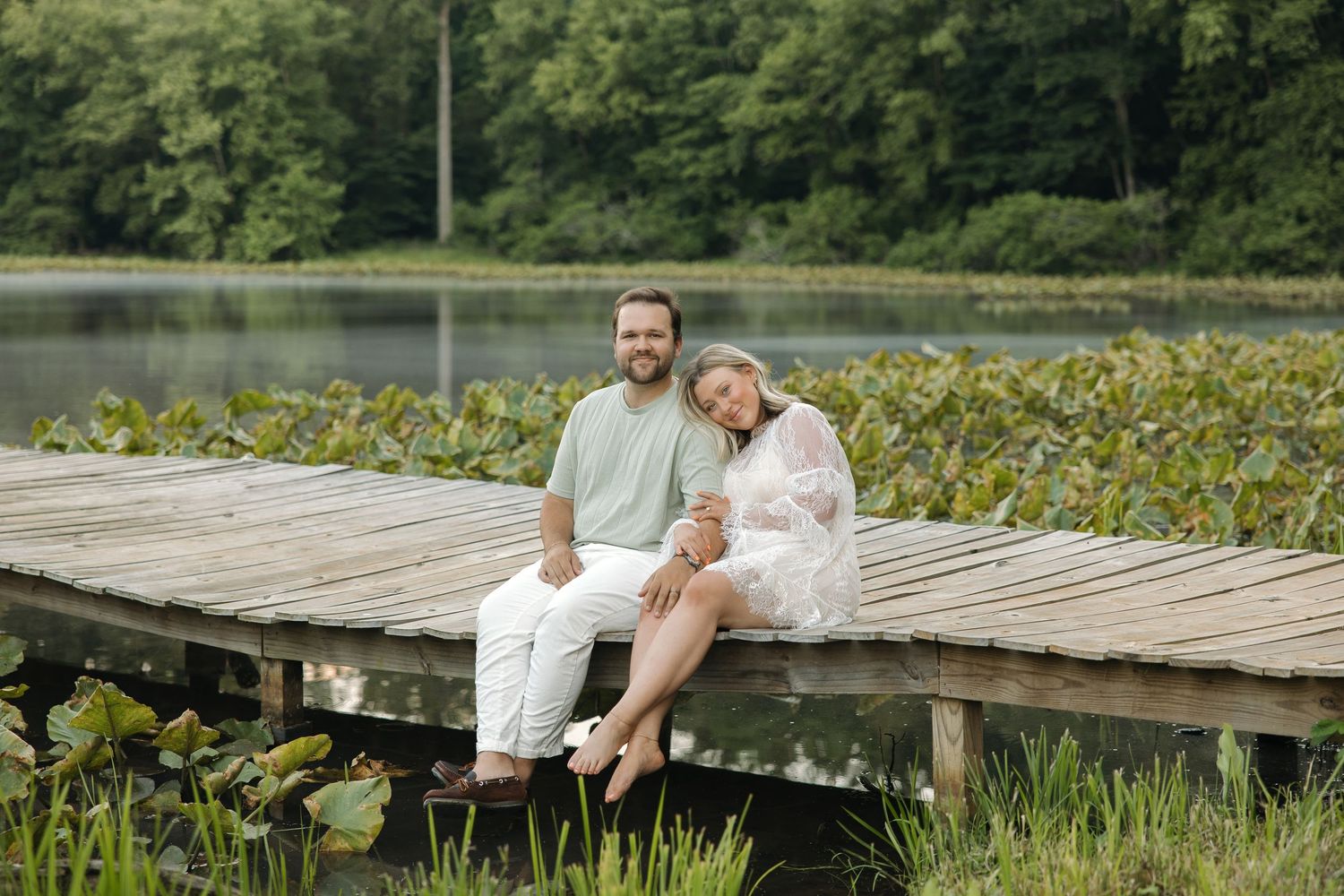 Smiling couple sitting together on a wooden lakeside dock surrounded by lily pads and green forest.