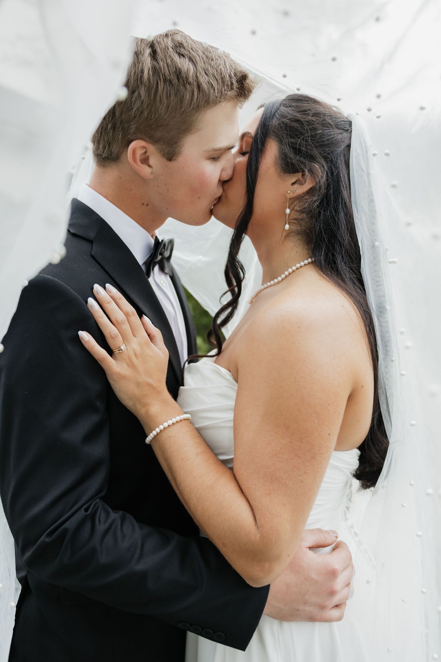 Bride and groom share a tender kiss beneath a pearl-adorned wedding veil, bride wearing pearl bracelet.