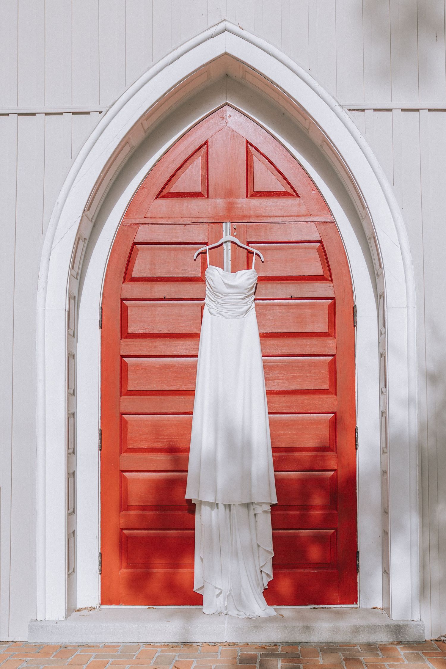 Elegant white wedding dress hanging in Gothic arched red church door entrance.