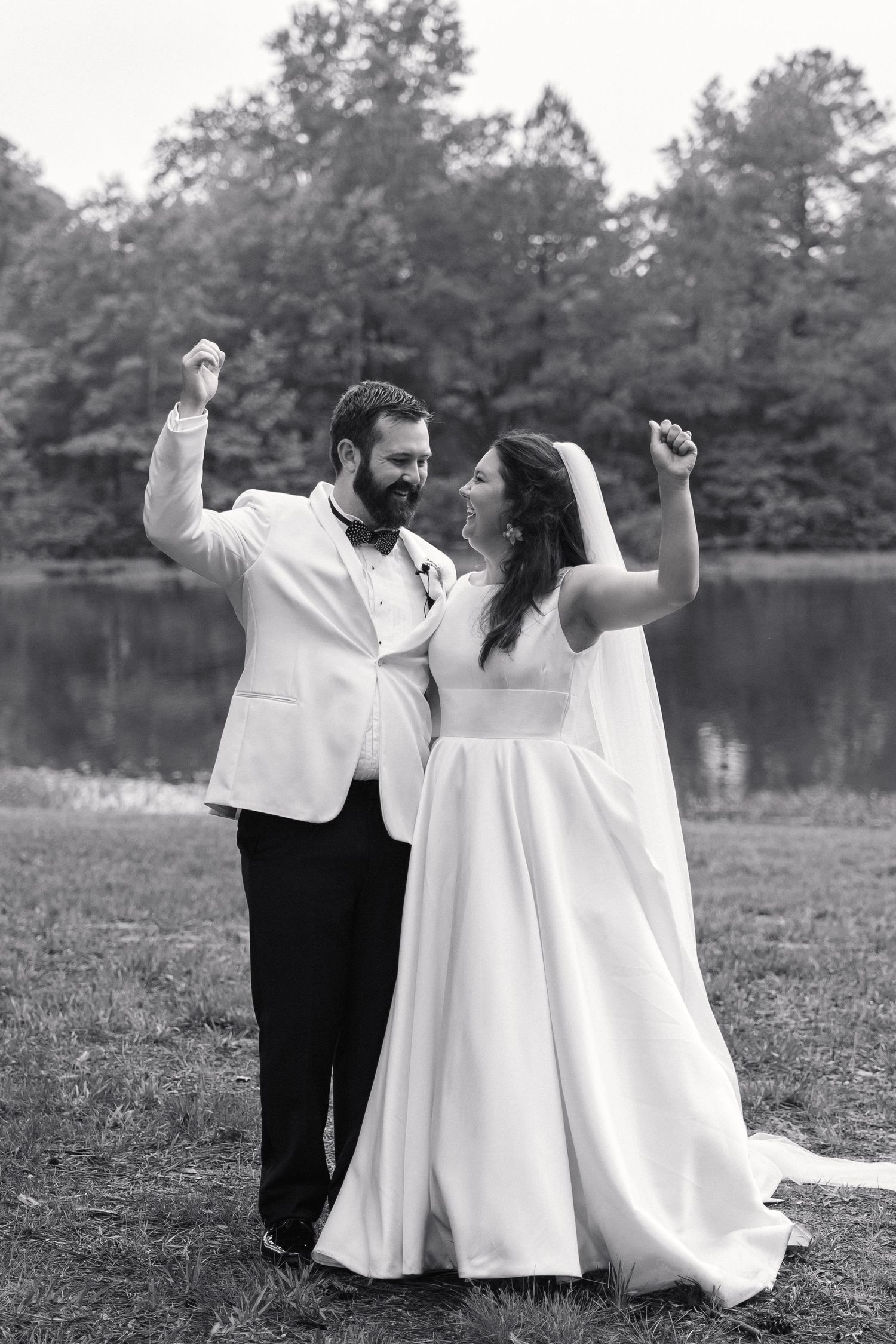 Joyful bride and groom celebrating with raised fists by a lake in black and white wedding photo.