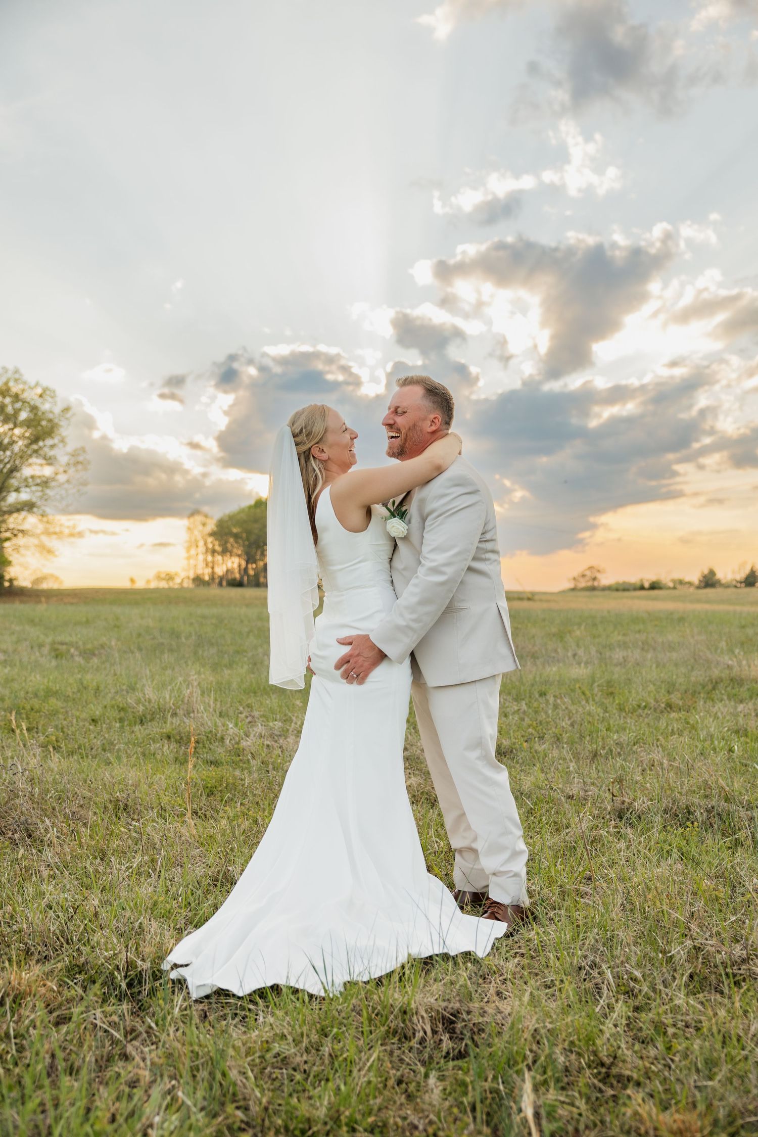 Bride and groom laugh and embrace in an open field beneath a dramatic cloudy sunset sky.