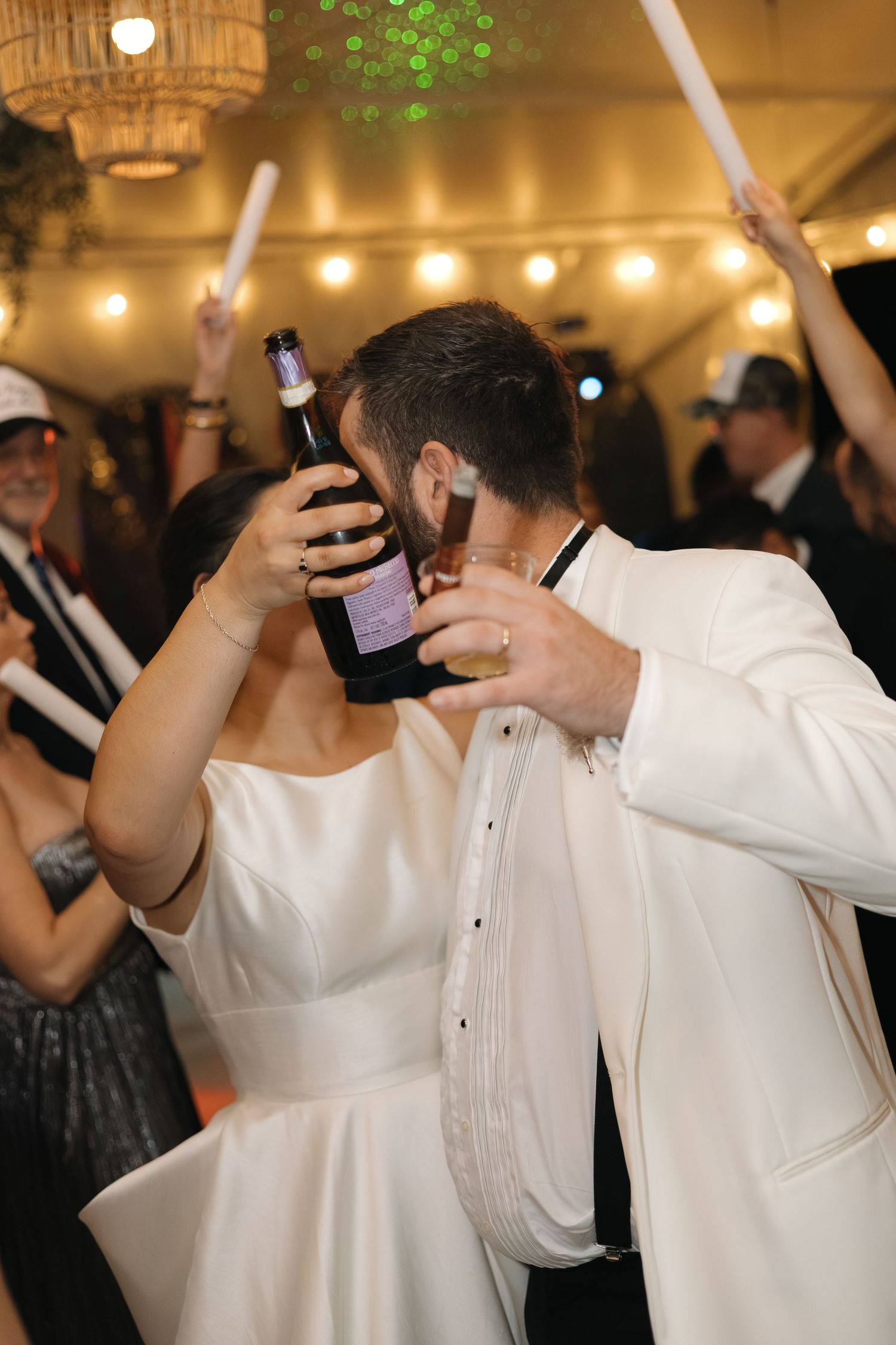 Bride and groom sharing a kiss while holding a wine bottle on the wedding dance floor surrounded by guests with glow sticks.
