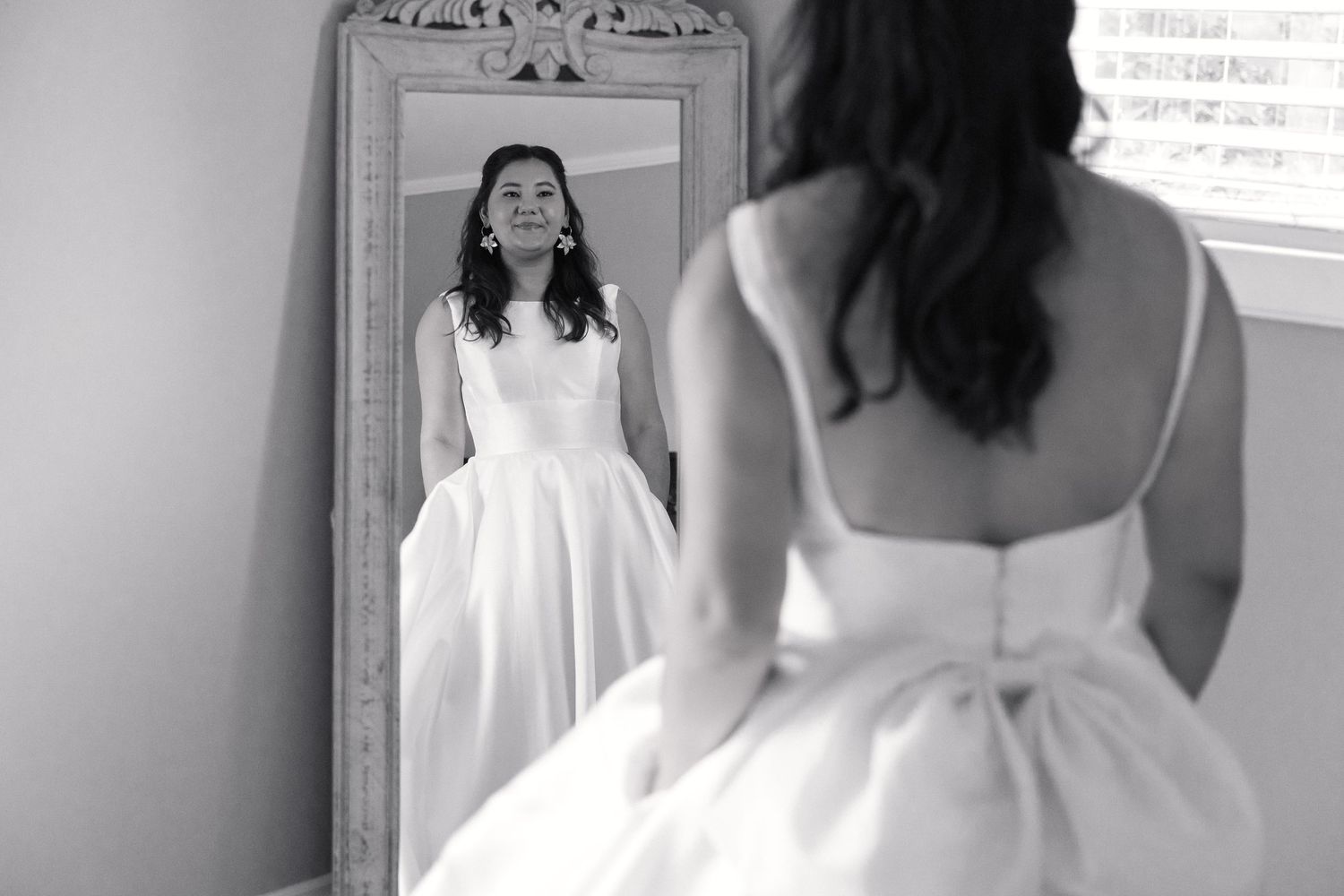 Bride in white ball gown admires her reflection in an ornate vintage mirror in black and white photo.