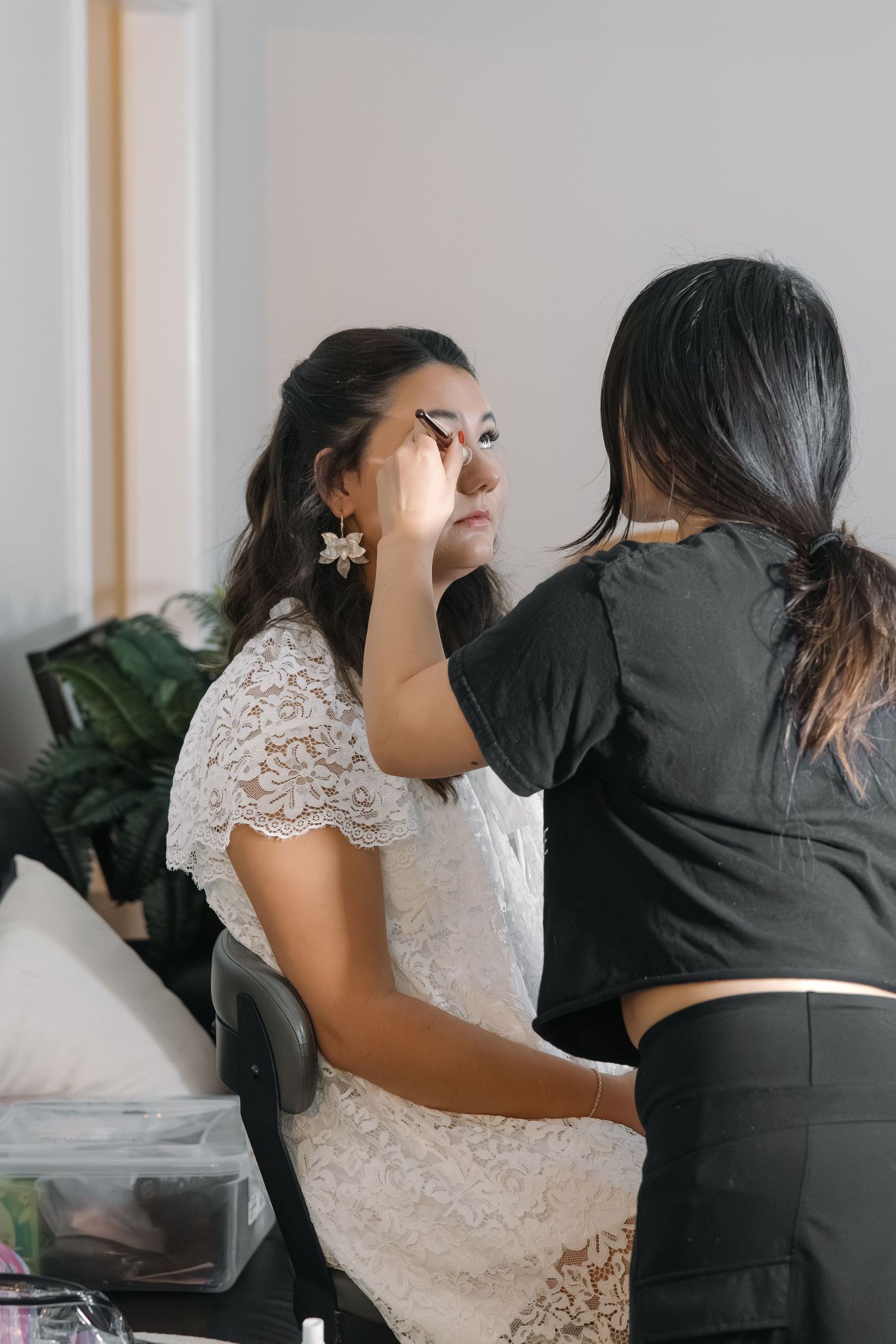 Professional makeup artist applying eye makeup to bride wearing white lace top before wedding.