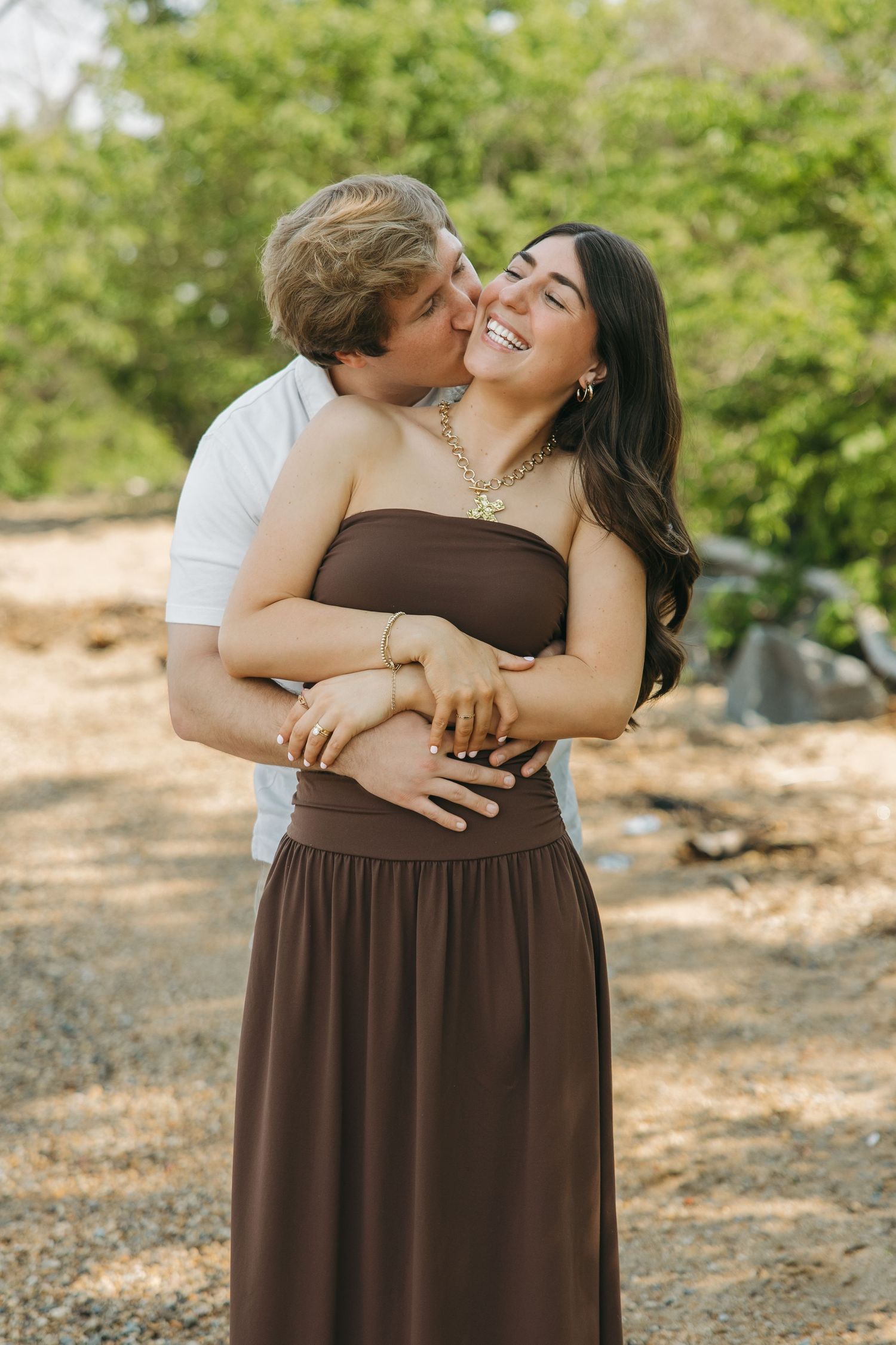 Man kissing woman on the cheek while embracing her from behind outdoors in nature.