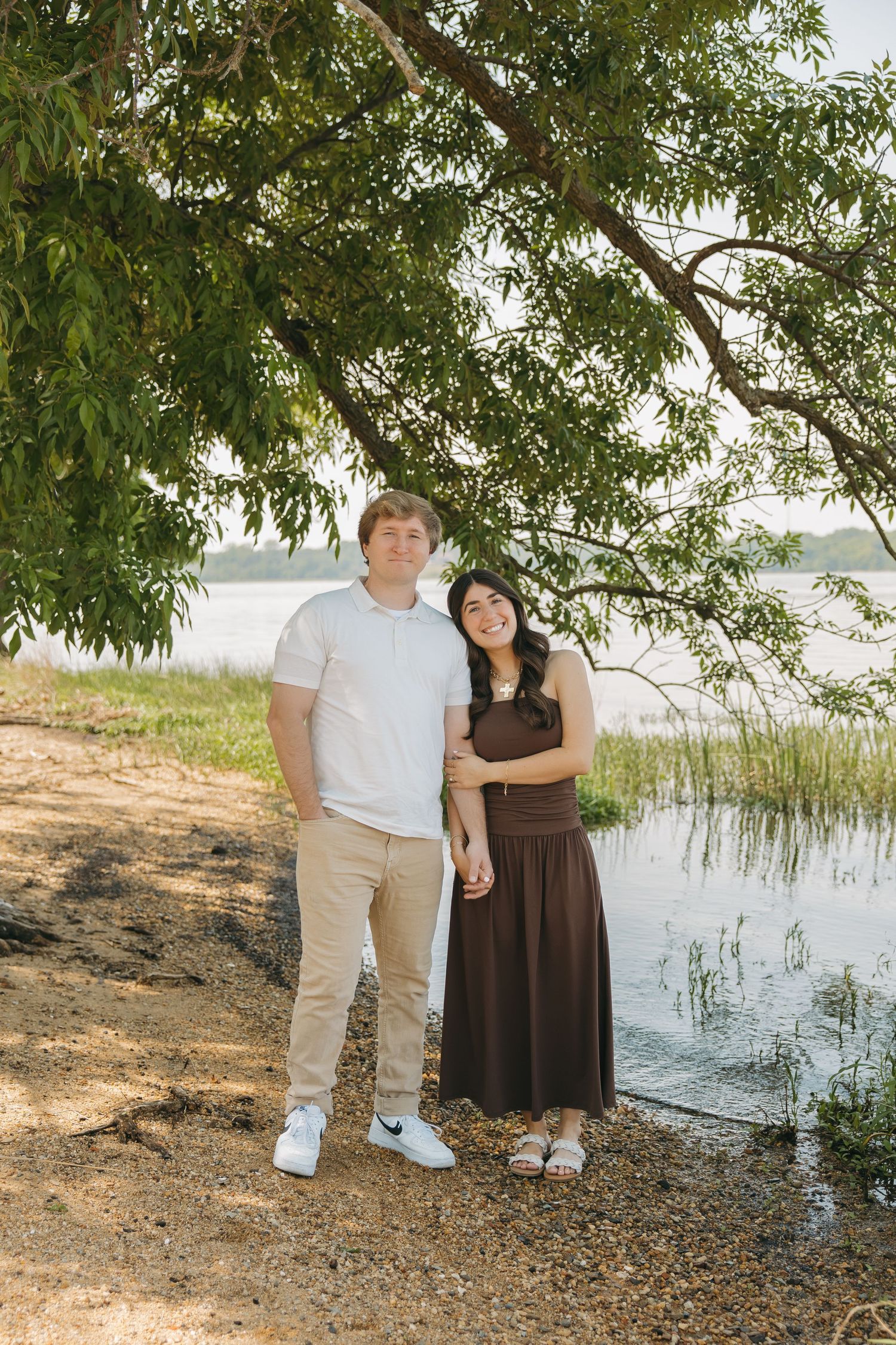 A couple stands together holding hands near a calm lake under a large overhanging tree outdoors.