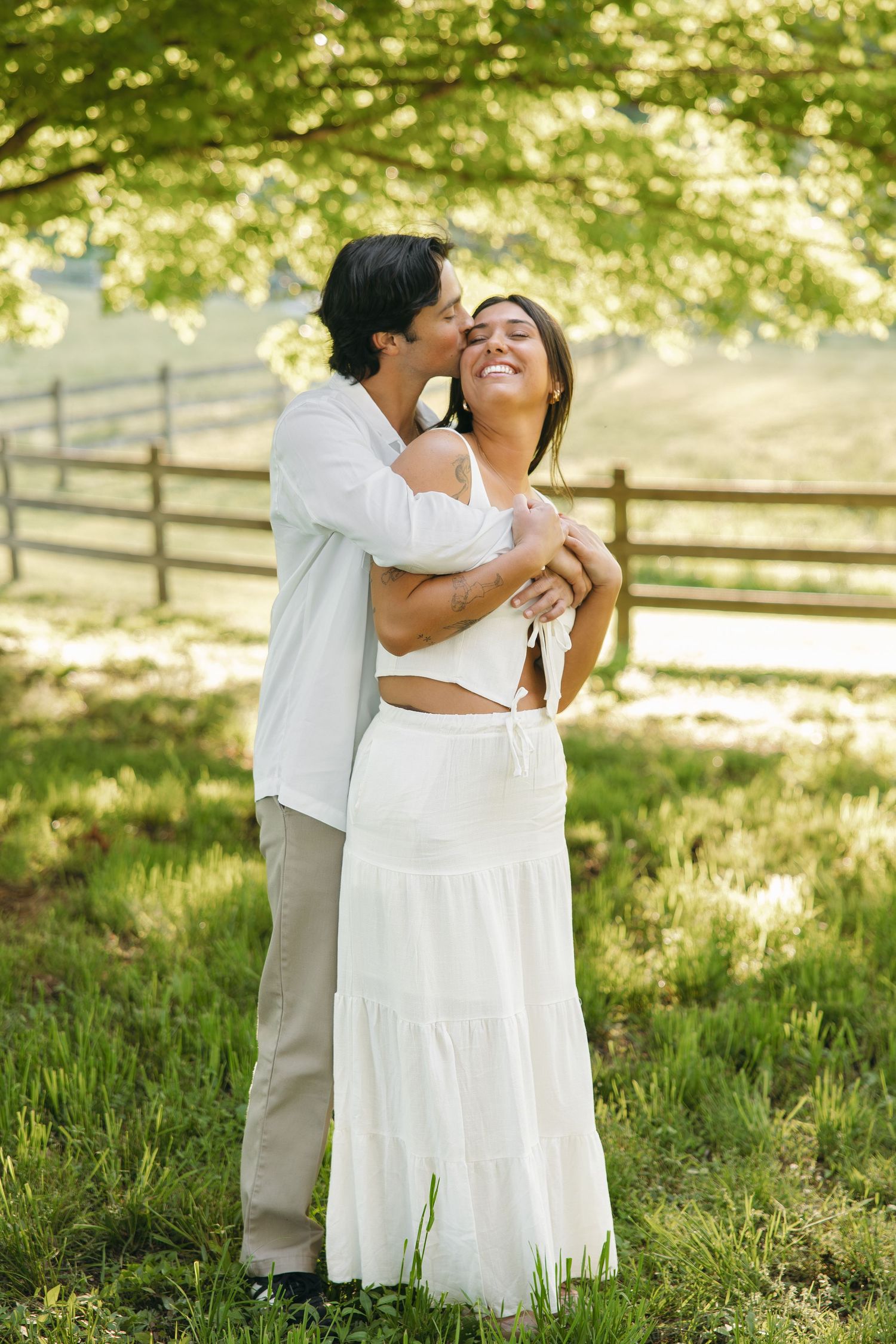 Happy couple embracing in sunlit park, man kissing smiling woman's cheek, both dressed in white near rustic fence.