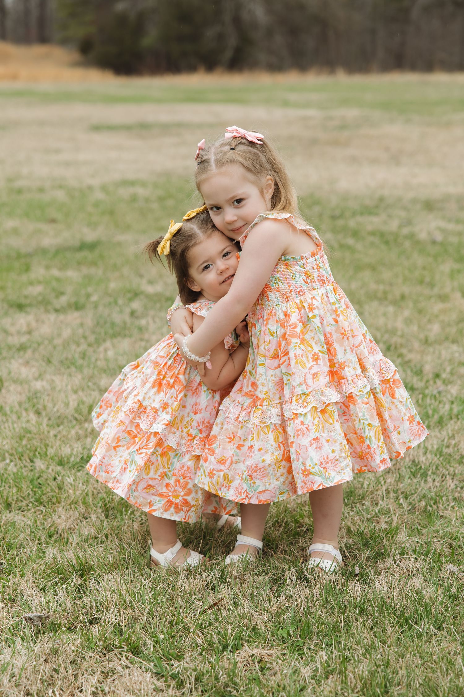 Two young sisters hugging in a grassy field, both wearing matching orange floral ruffle dresses with hair accessories.