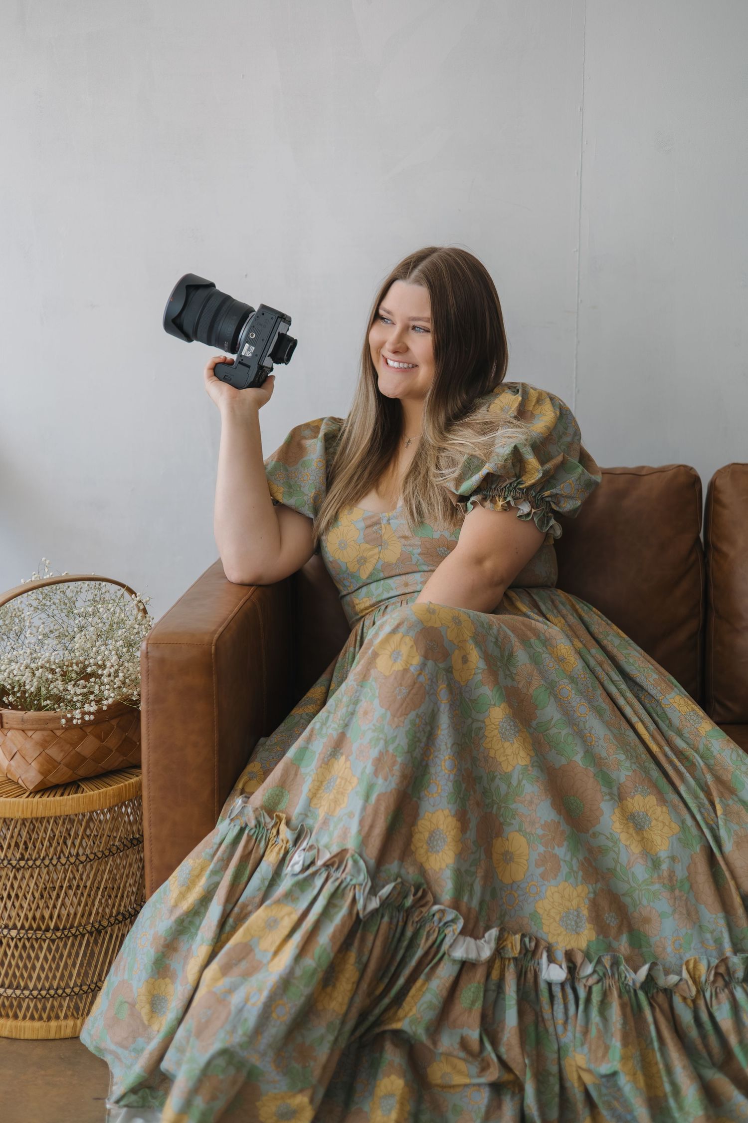 Smiling female photographer in floral dress holding camera while seated on brown leather sofa.
