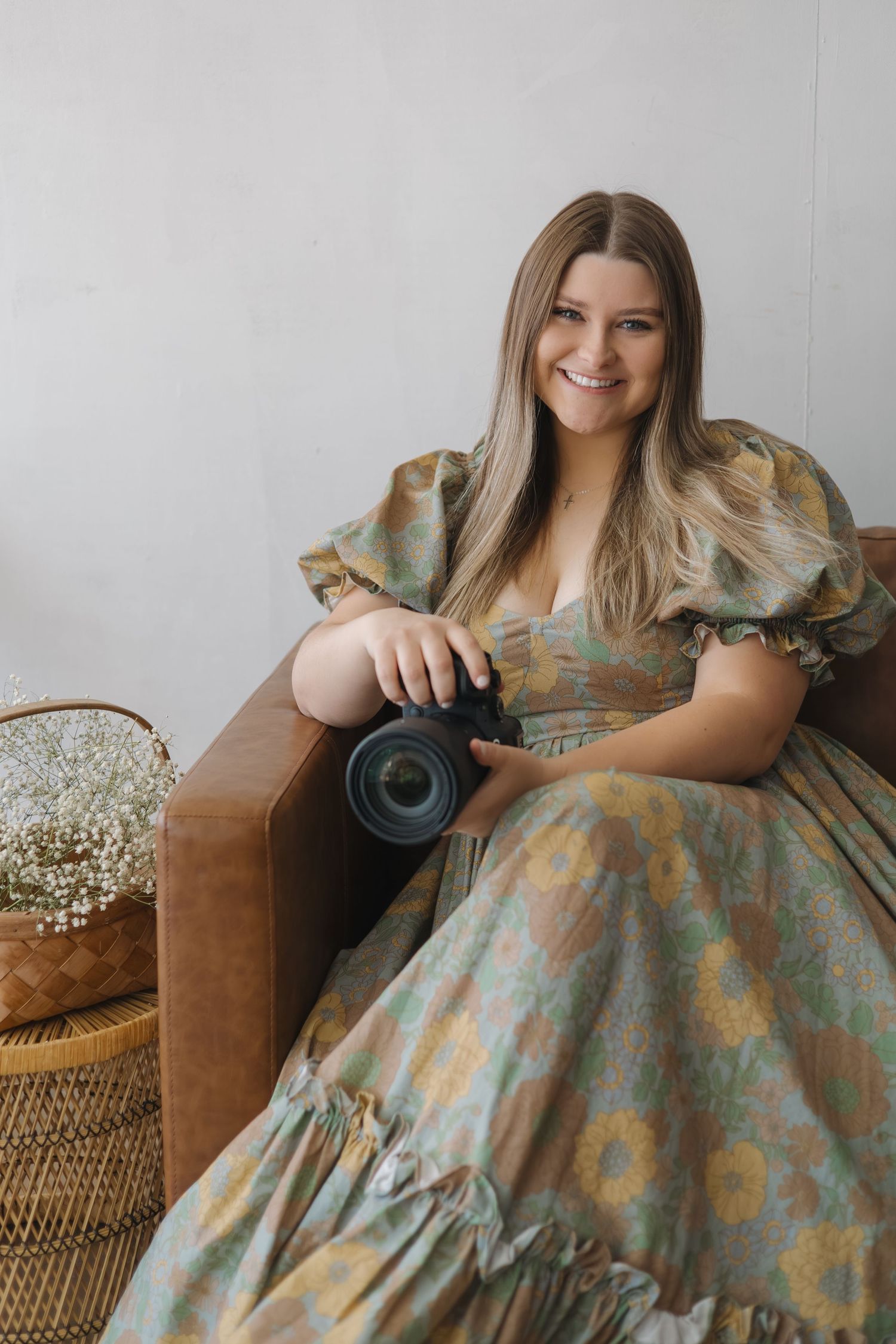 Smiling woman in floral dress holding a camera while seated on a leather chair with baby's breath flowers nearby.