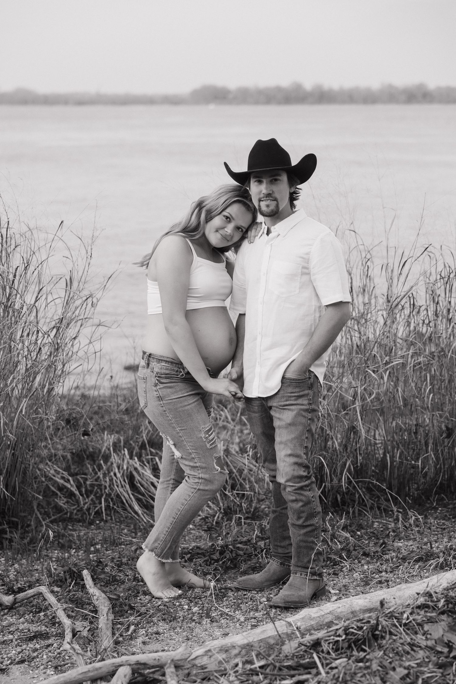 Black and white maternity portrait of pregnant couple standing by tall reeds near water, man wearing cowboy hat.