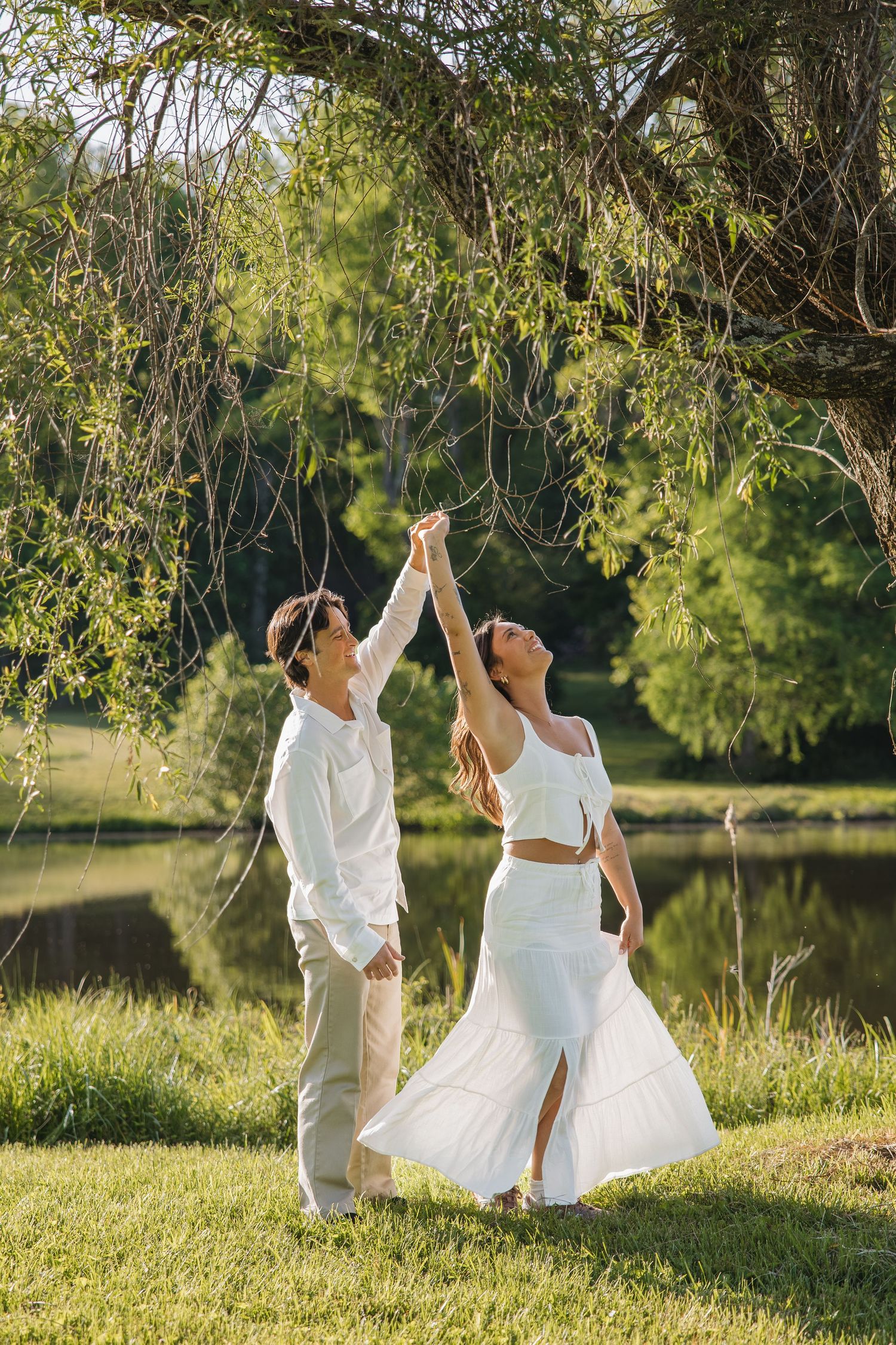 Happy couple holding hands and reaching toward weeping willow branches near a peaceful lake on a sunny day.
