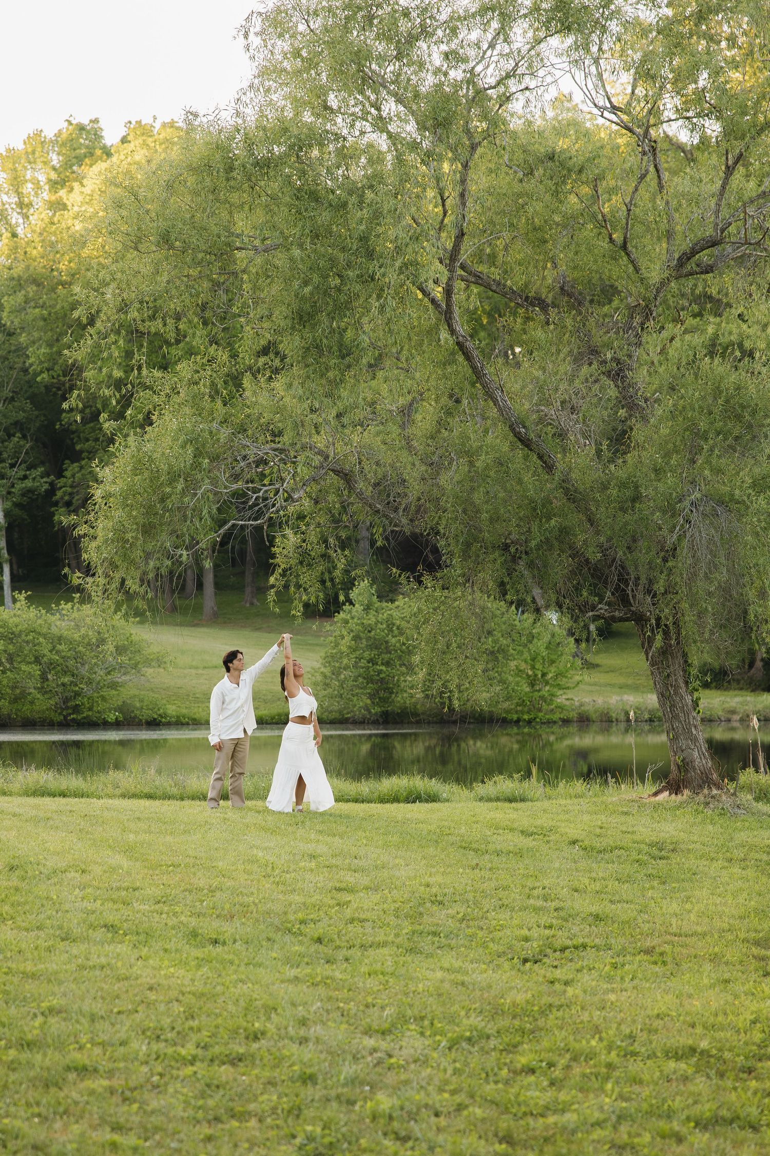 Couple holding hands and dancing near a large willow tree beside a tranquil pond in a park.