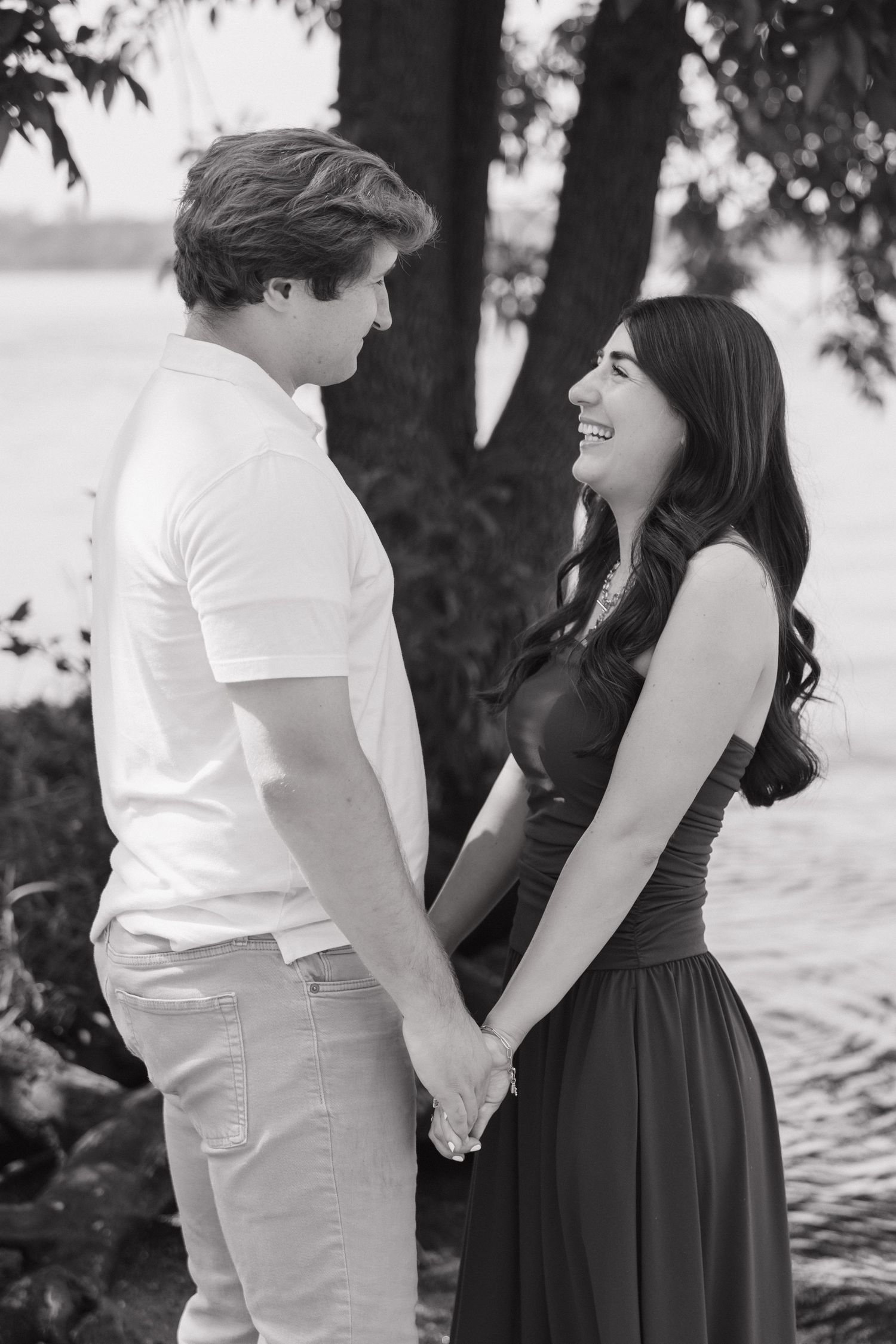 Black and white photo of a young couple holding hands and smiling by a lake.