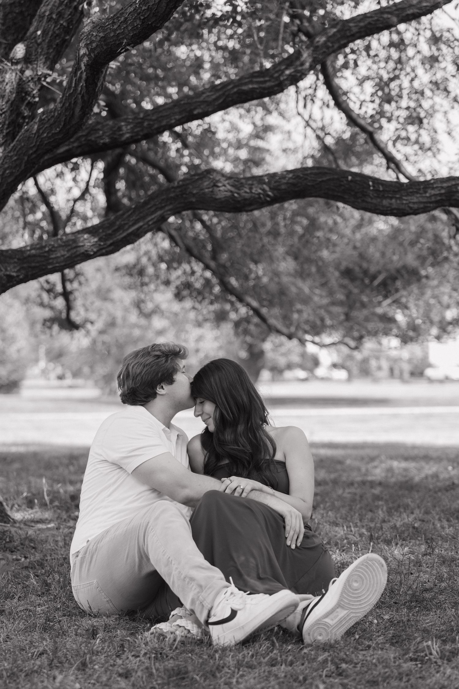Black and white photo of a couple sitting on grass under a tree, leaning in close to each other.