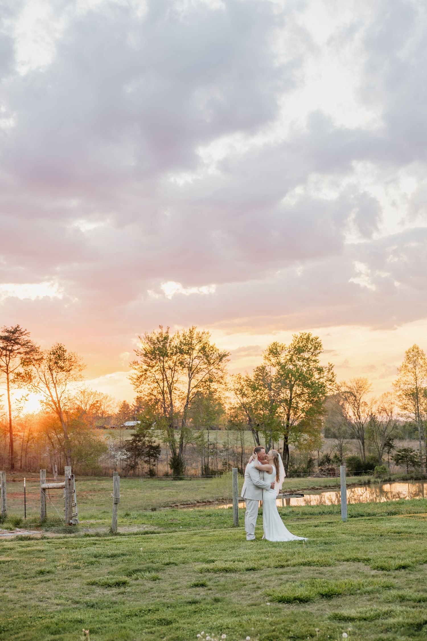 Bride and groom kissing in an open green field under a dramatic colorful sunset sky.