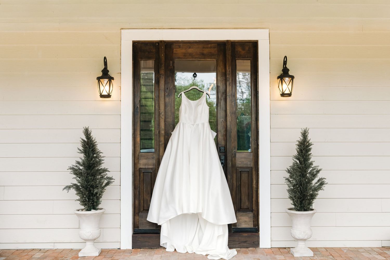 Elegant white ball gown wedding dress hanging on a rustic wooden front door flanked by potted evergreen trees.