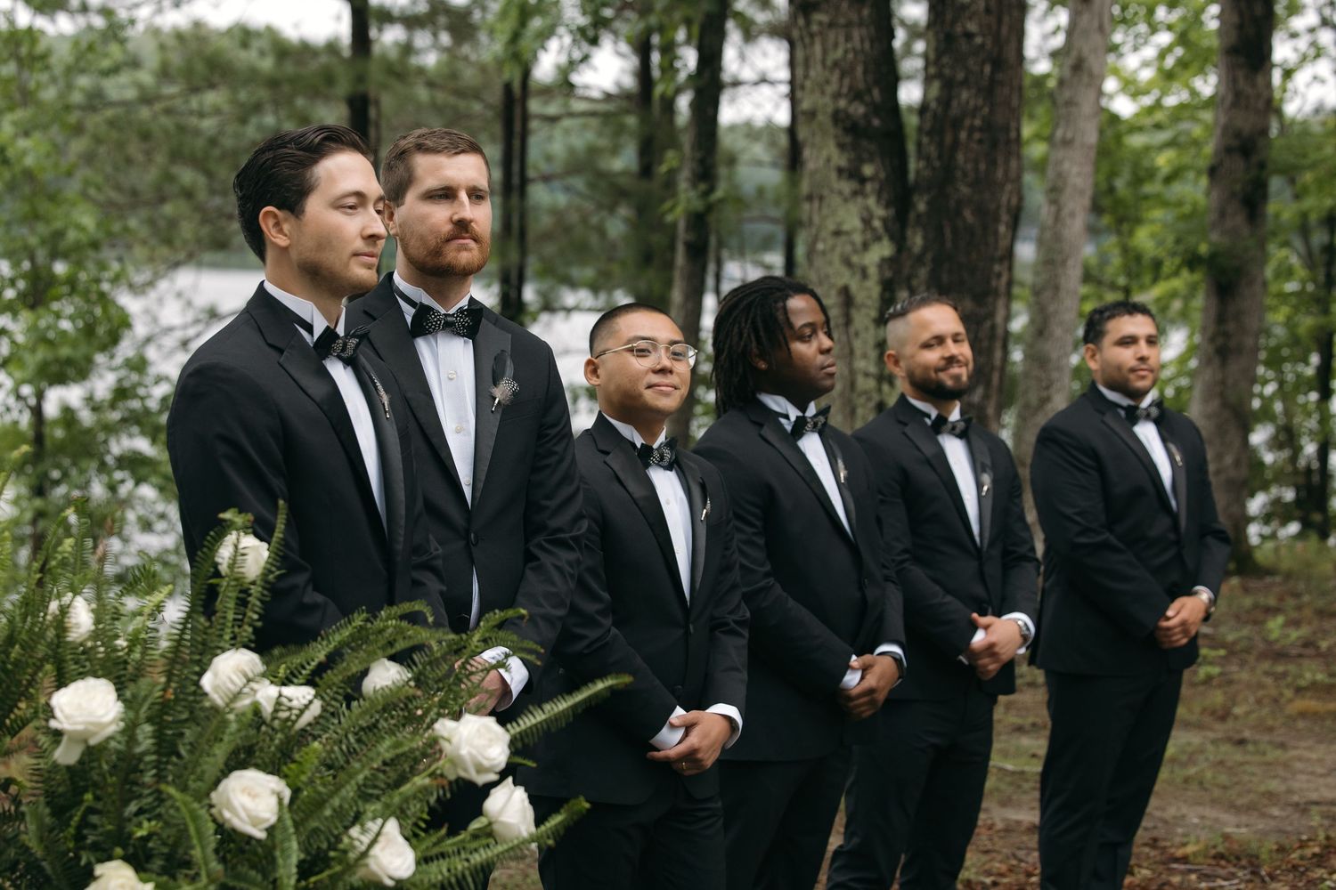 Six groomsmen in black tuxedos standing at attention during outdoor lakeside wedding ceremony.