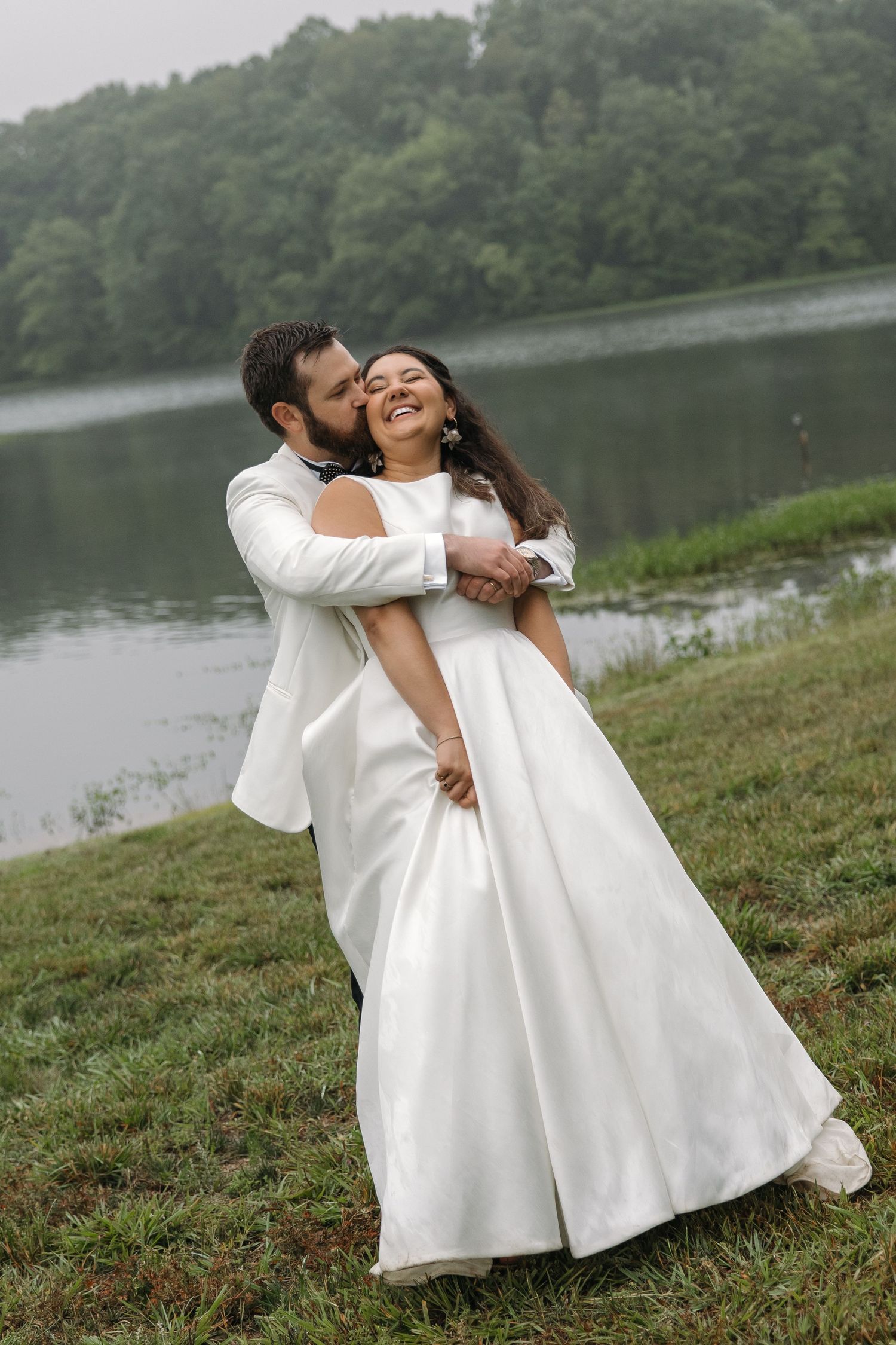 Groom in white tuxedo embraces bride in elegant white gown by misty lakeside.