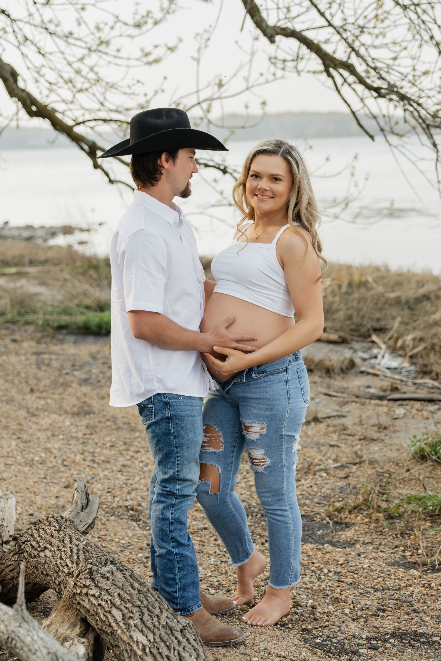 Couple holding pregnant belly outdoors near water, man in black cowboy hat and white shirt, woman in ripped jeans.
