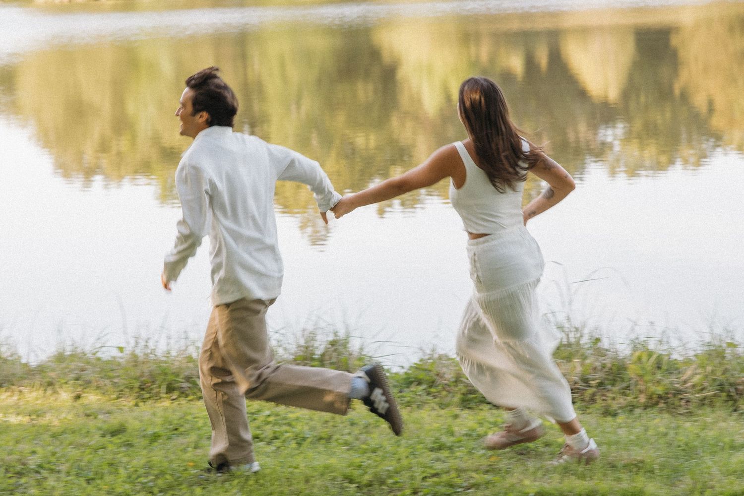 Couple running hand-in-hand along a grassy lakeside in casual white outfits during golden hour.