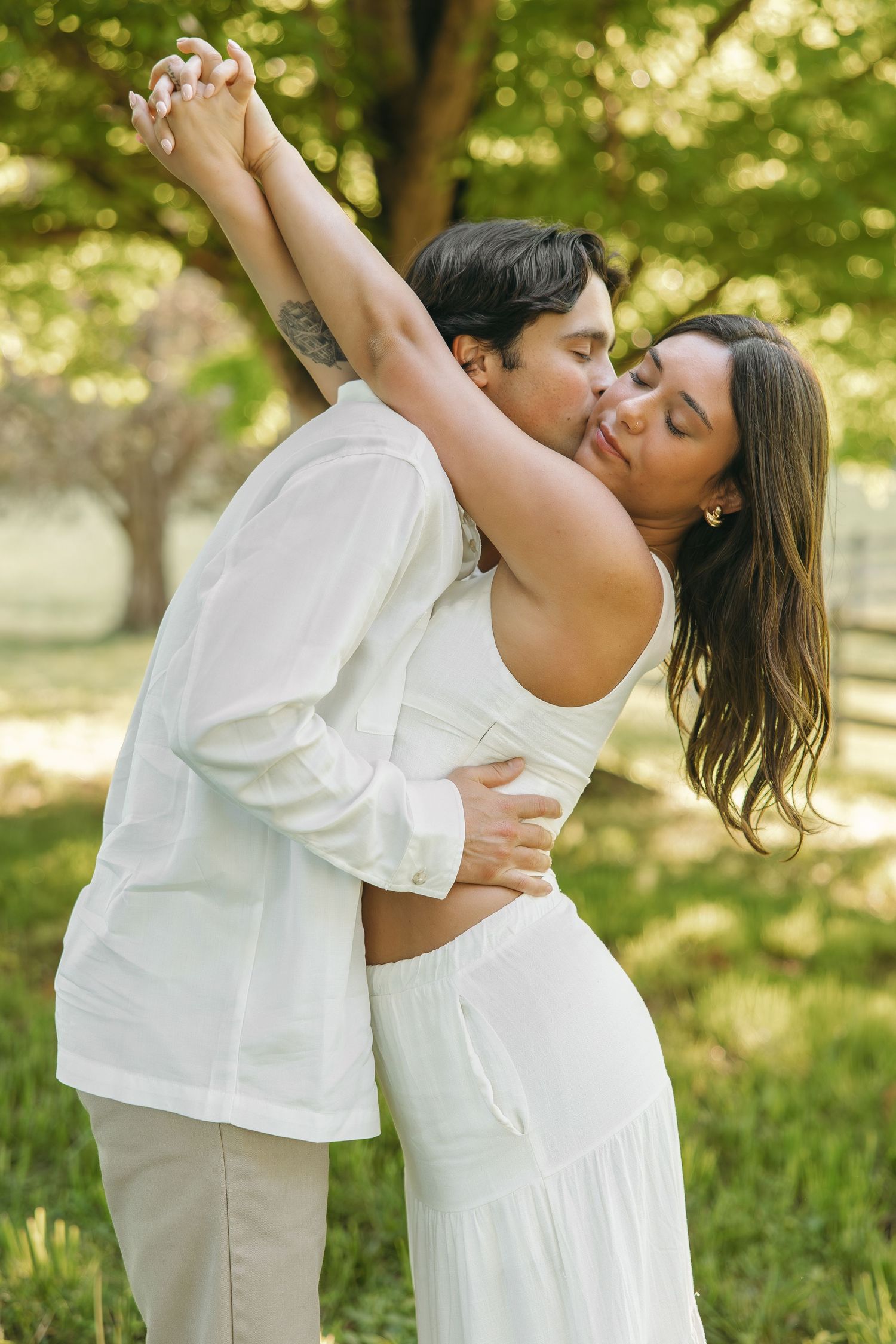Couple dancing romantically outdoors, holding hands raised while sharing a kiss, surrounded by green trees and nature.