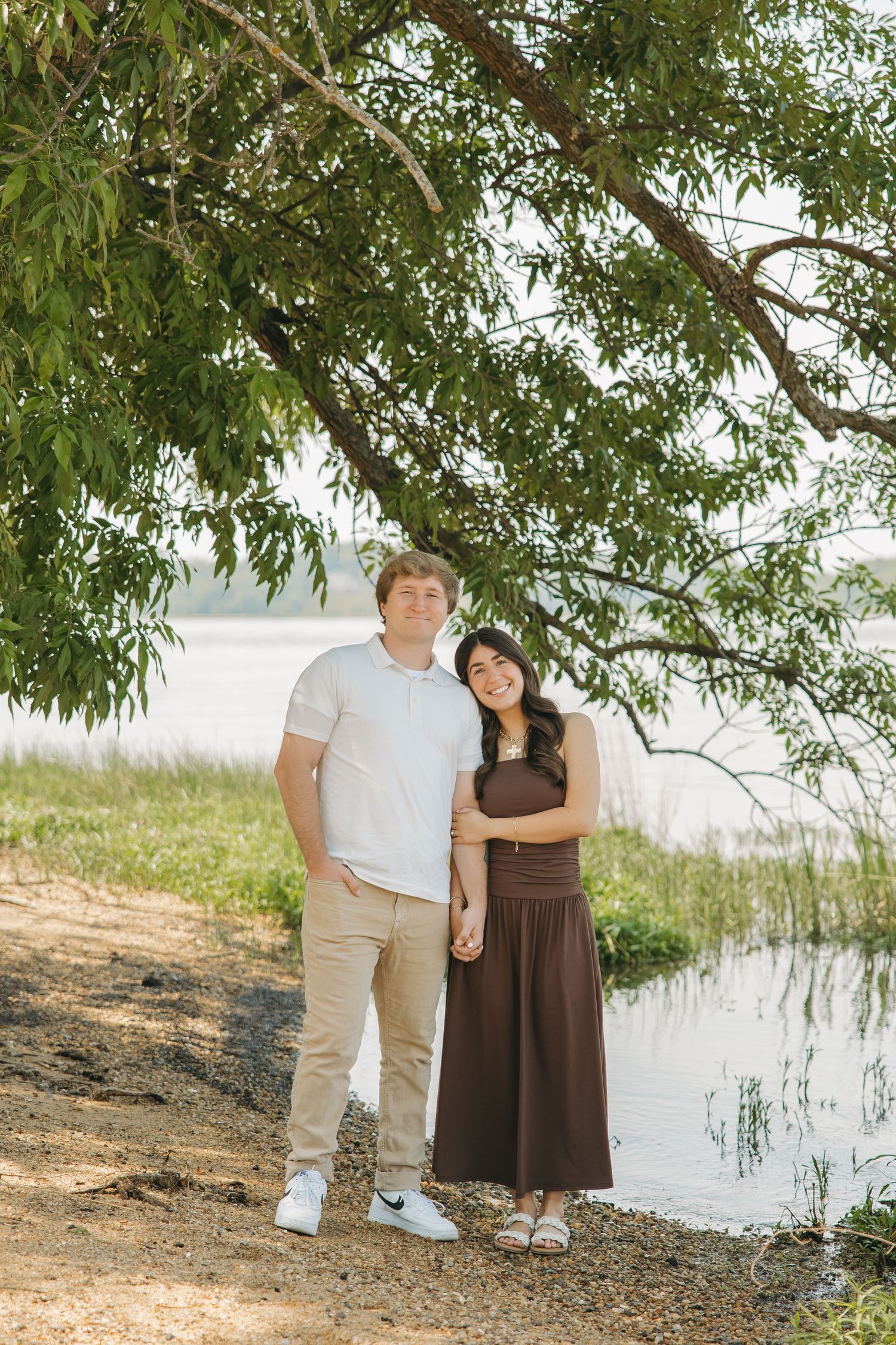 Young couple holding hands and standing under a willow tree by a calm lakeside.