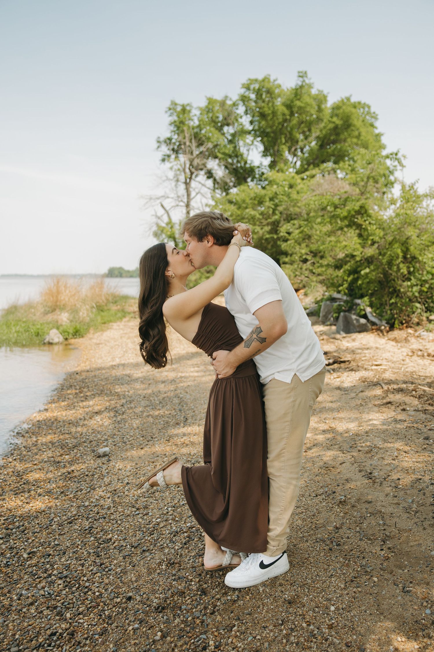 A romantic couple kisses near a lakeside, woman in brown maxi dress lifting her foot as they embrace.