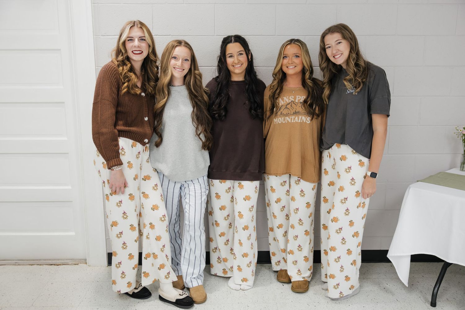 Five women pose together smiling in matching floral pajama pants and cozy fall-toned tops.