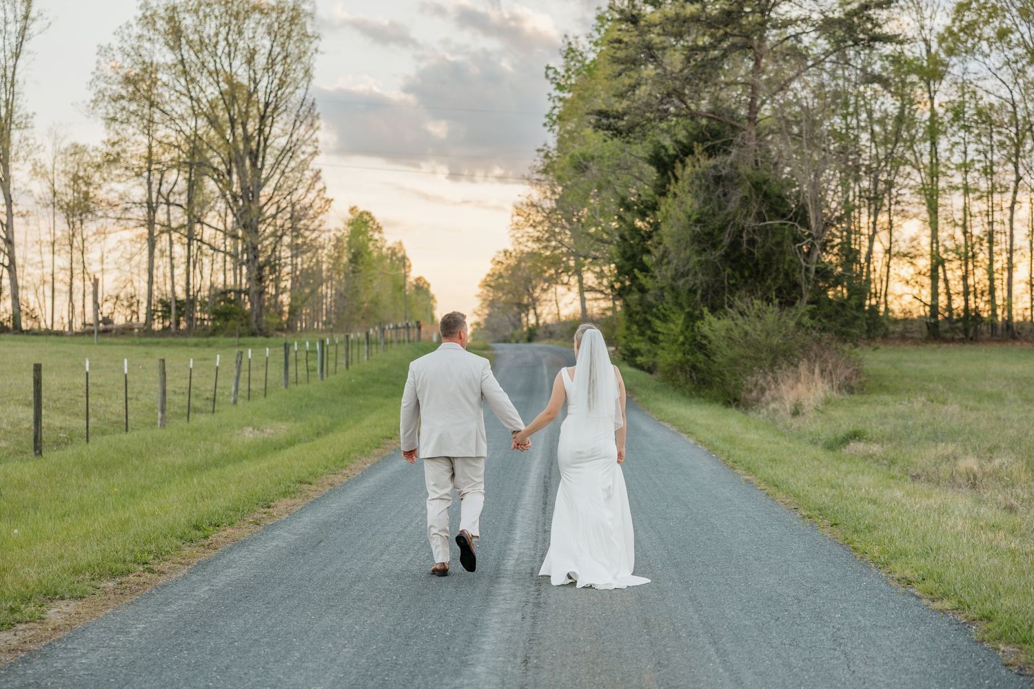 Bride and groom walking hand in hand down a rural road at sunset with green fields on either side.