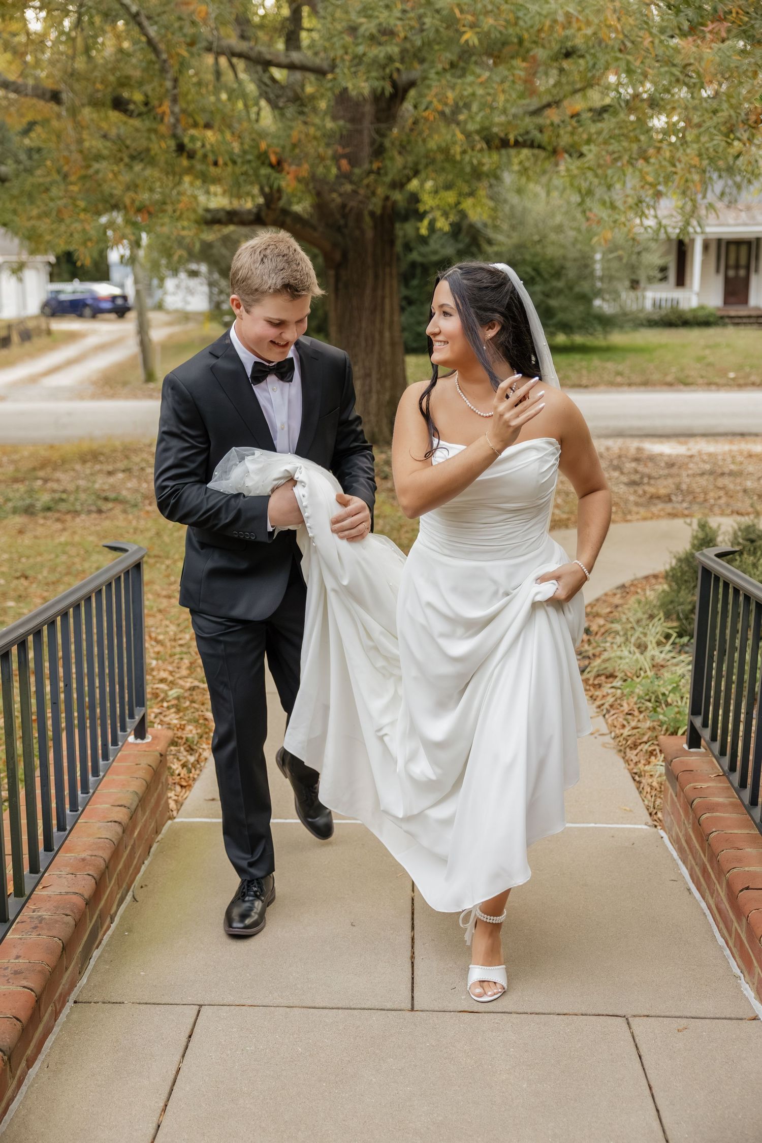 Groom in black tuxedo helping bride in white strapless gown carry her dress train on a walkway outdoors.