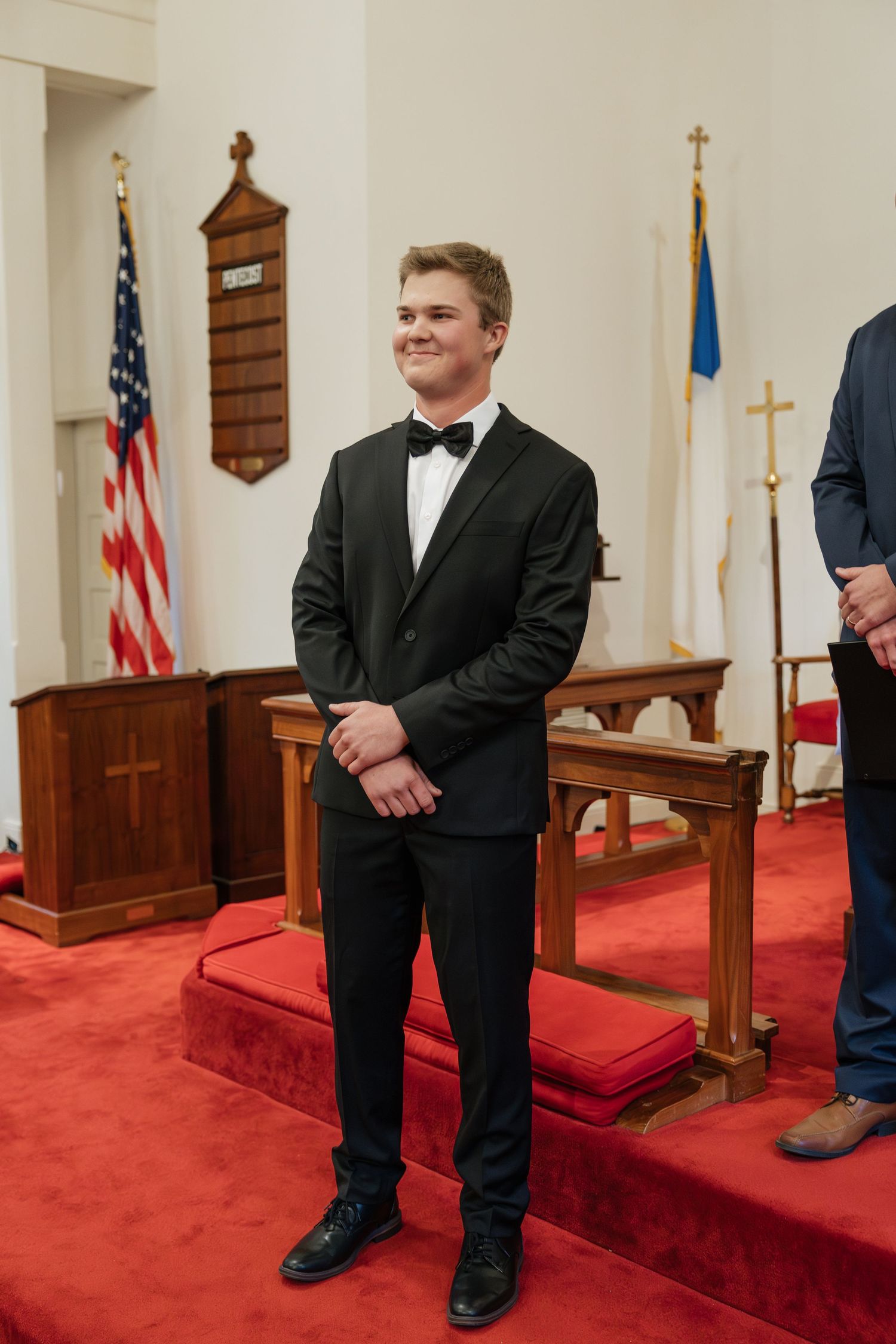 Smiling groom in black tuxedo with bow tie standing at the altar of a church with red carpet.