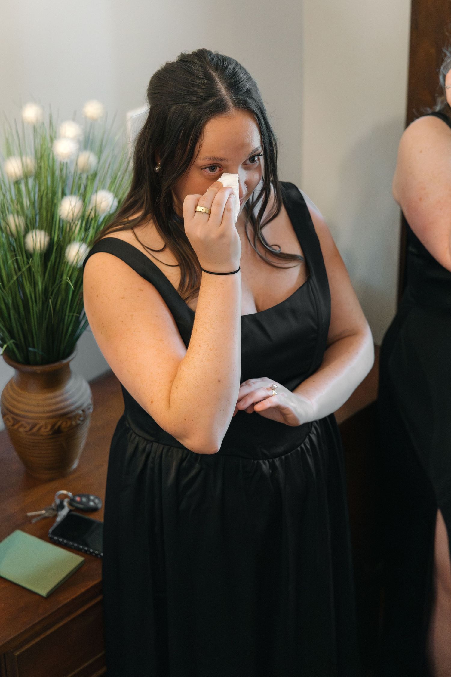 Emotional bridesmaid in black dress dabs tears with tissue during wedding day preparations.